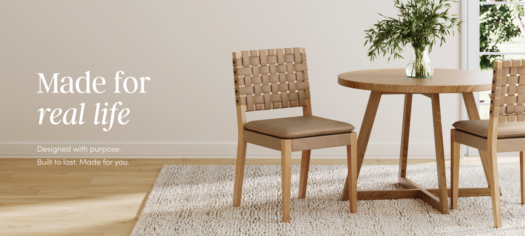 A minimalist dining area with a round wooden table, two woven-back wooden chairs, a textured rug, and a green plant in a vase. Text reads: Made for real life. Designed with purpose. Built to last. Made for you.