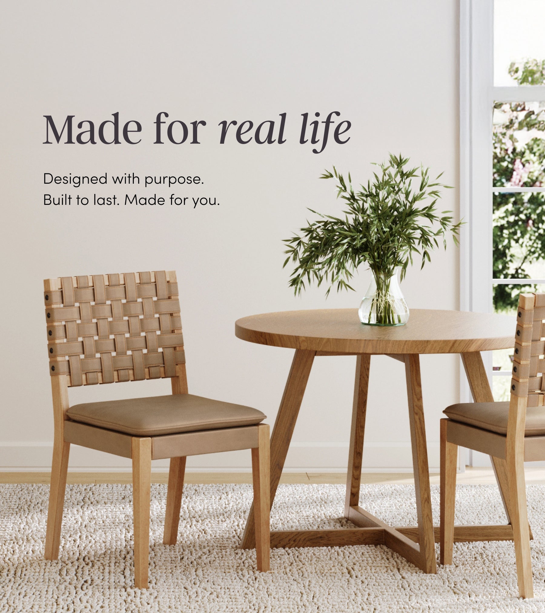 A modern dining area with a round wooden table, two woven-back chairs, a glass vase with green leafy branches, and sunlight streaming through a window. Text on the wall reads, Made for real life.