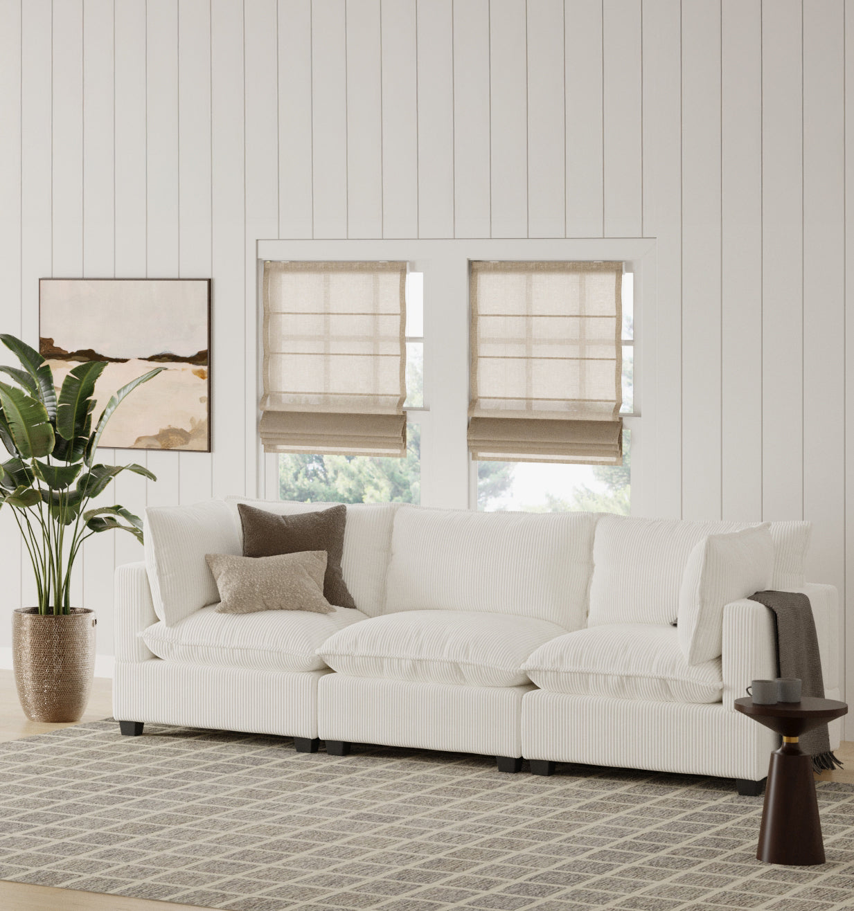 A modern living room with a large white sectional sofa, neutral throw pillows, a brown blanket, a potted plant, a round side table, a geometric rug, wall art, and two windows with beige Roman shades.