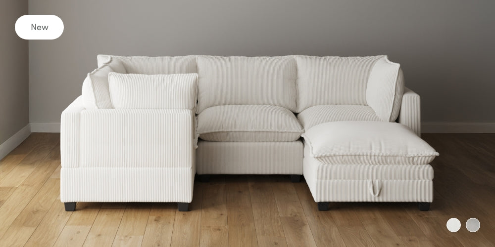 A modern white sectional sofa with ribbed fabric sits on a wooden floor against a plain gray wall. The sofa has plush cushions and a chaise lounge on the right side.