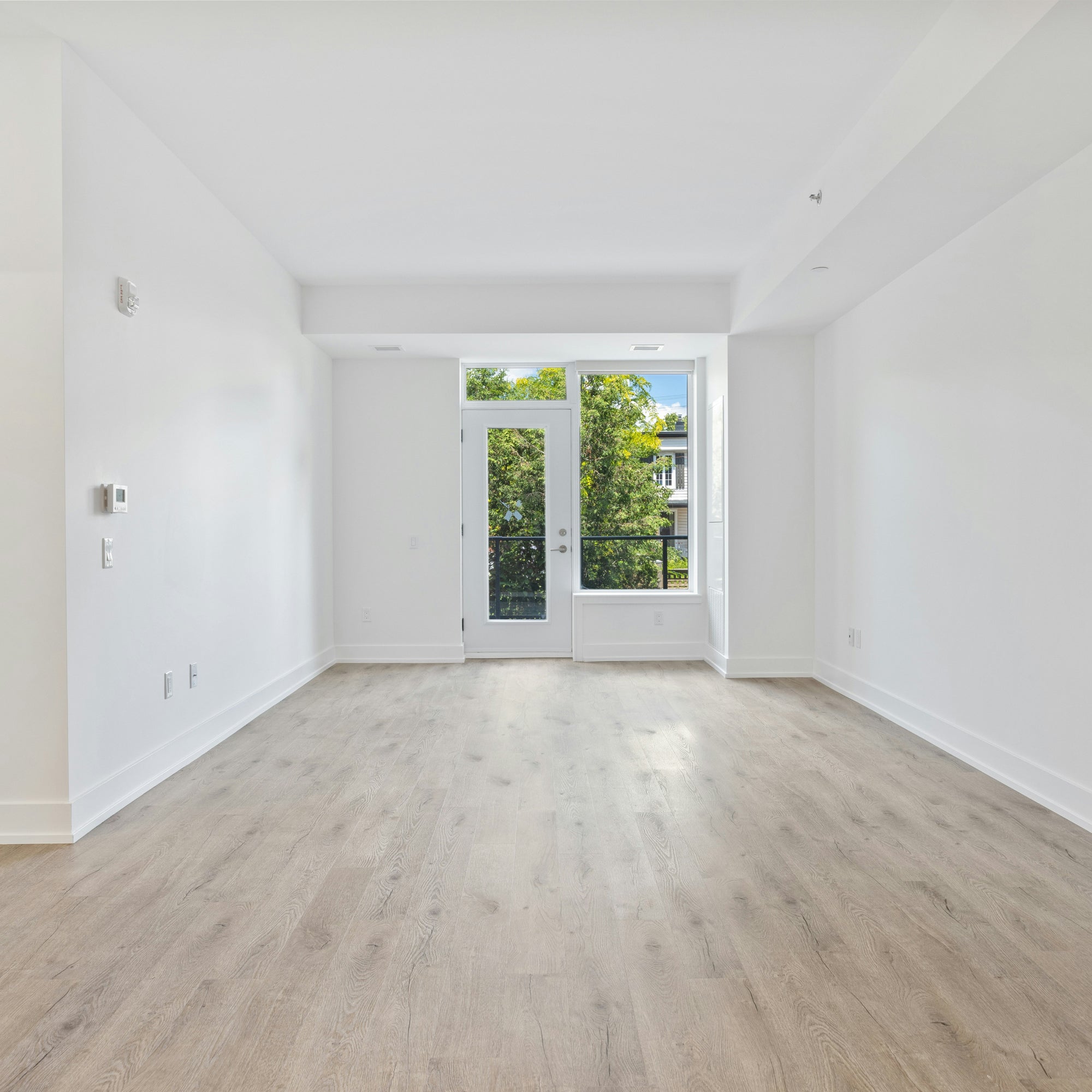 Bright, empty room with light wood flooring, white walls, and large glass doors leading to a balcony surrounded by greenery. Natural light fills the modern, minimalist space.