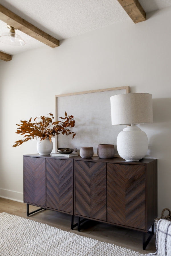 A modern wooden sideboard with a chevron pattern holds a white vase with brown leaves, two small brown pots, a white lamp, and a framed picture, set against a light wall in a cozy, neutral-toned room.