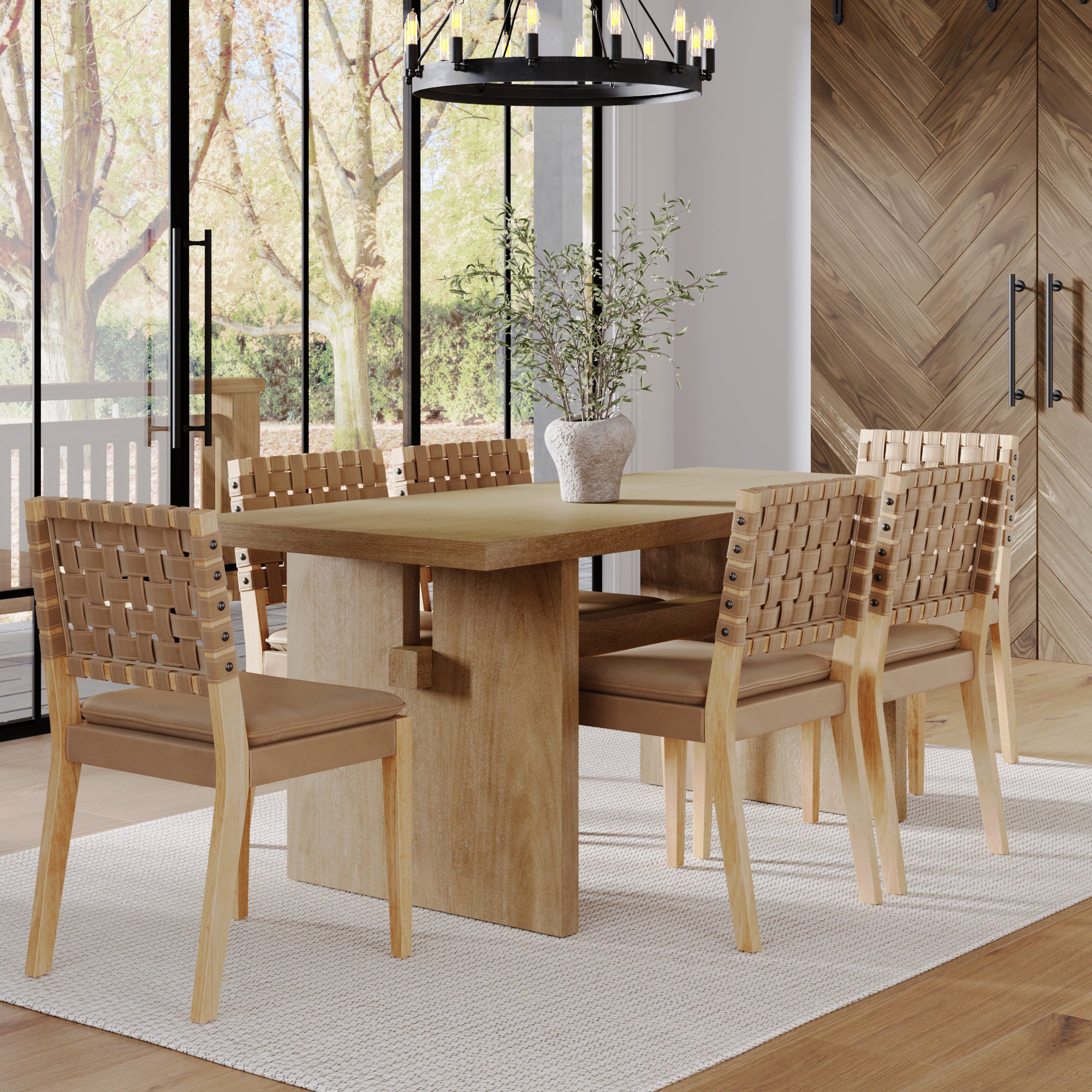 A modern dining room features the Nathan James Mid-Century Woven Dining Set in light brown with six chairs, a white vase with greenery, large windows overlooking trees, a black chandelier above, and a white rug below.