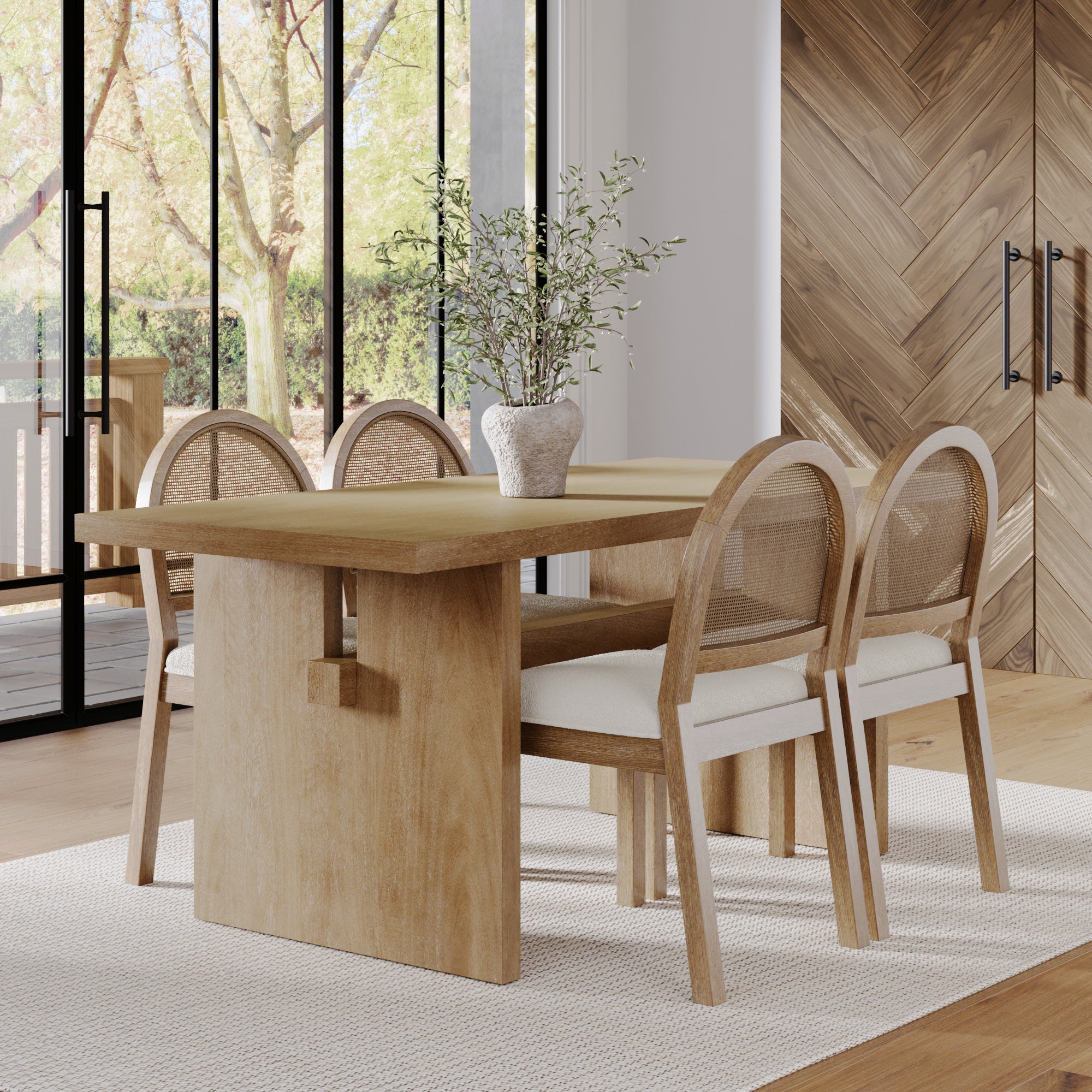 A modern dining room showcases the Nathan James Bohemian Cane Dining Set with a light wood veneer table and four rattan chairs with white cushions. A white vase with greenery decorates the table, while large windows reveal views of trees outside.