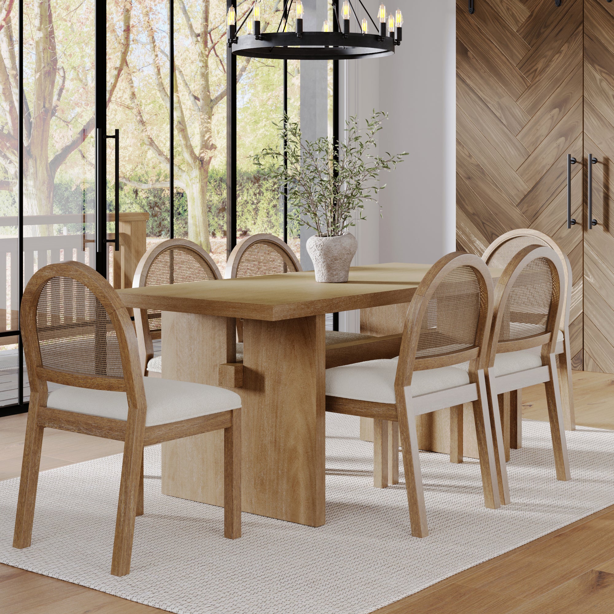 A modern dining room with the Nathan James Bohemian Cane Dining Set—solid wood veneer table, six cushioned rattan chairs—a potted plant centerpiece, large windows with tree views, a black chandelier above, all atop a light rug.