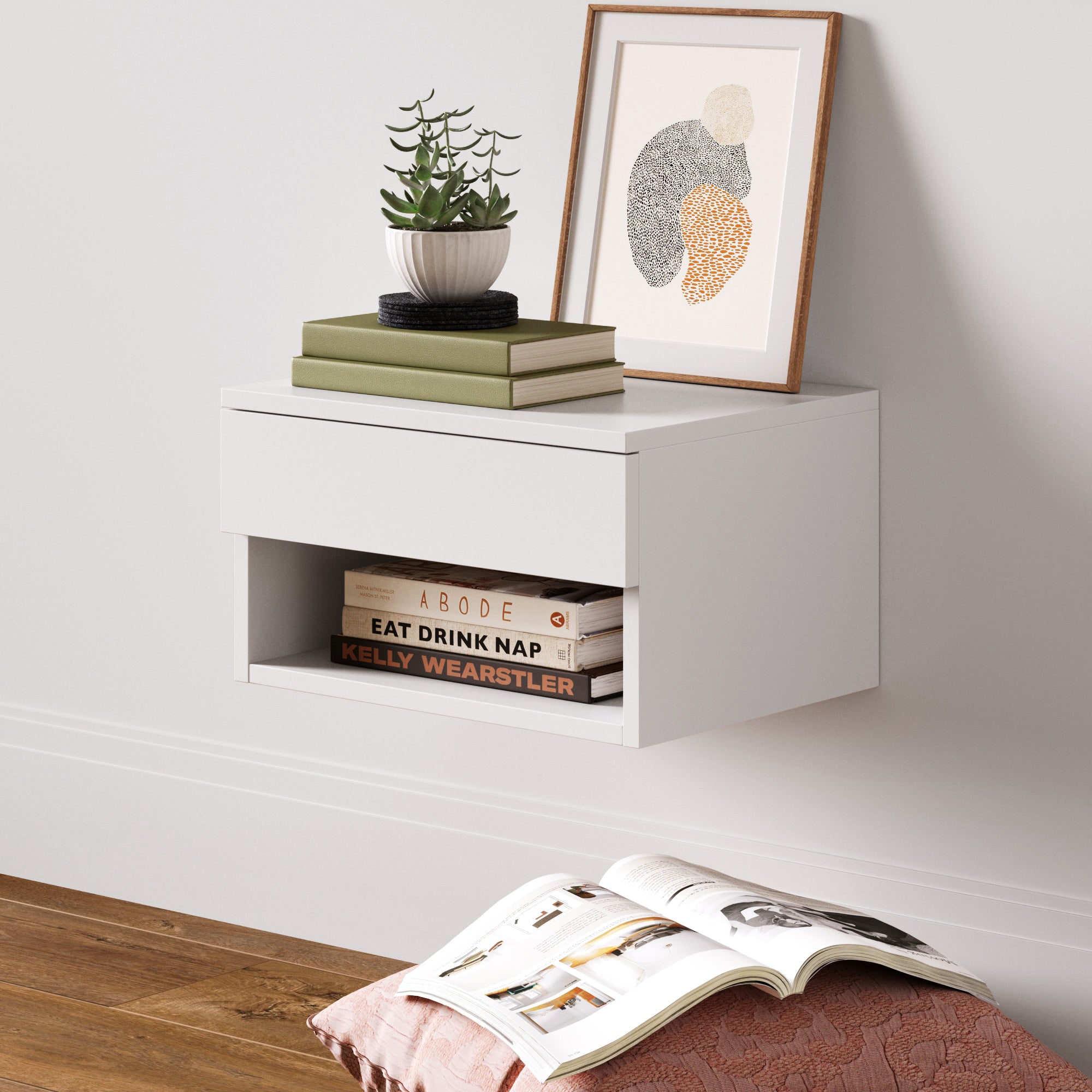 A modern white floating nightstand with a drawer and open shelf, holding books. On top are stacked books, a potted plant, and framed abstract art. A quilt and open magazine lie on the wooden floor nearby.