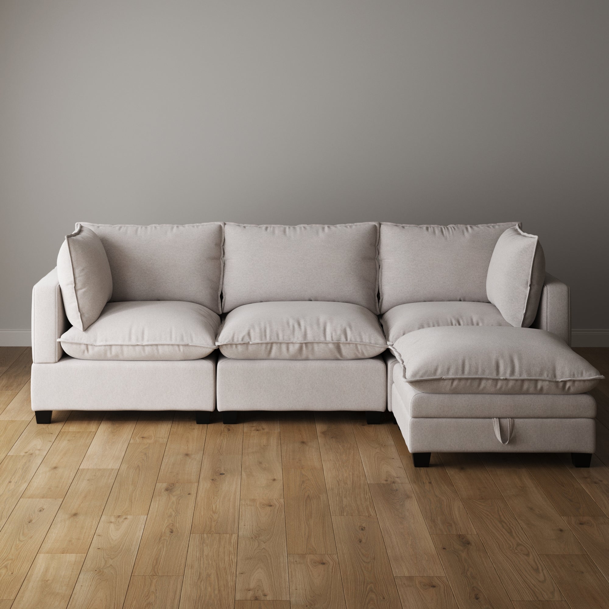 A modern, light gray sectional sofa with plush cushions is set against a plain light gray wall on a wooden floor in a minimalistic living room.