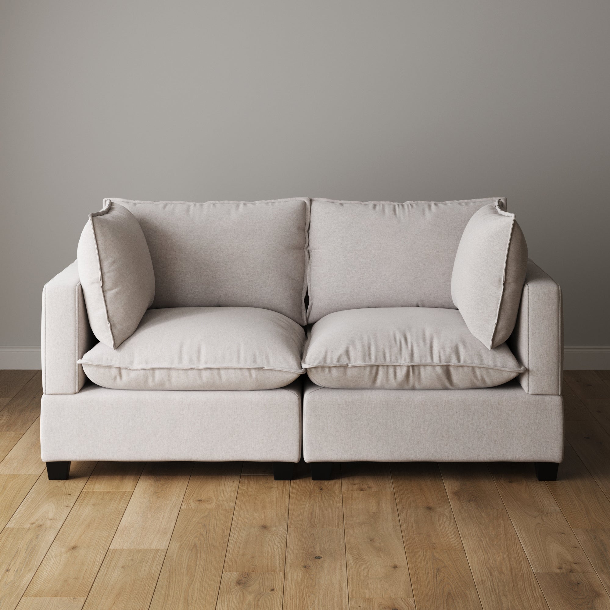 A light beige, modern two-seat sofa with plush cushions sits on a wooden floor against a plain light gray wall.