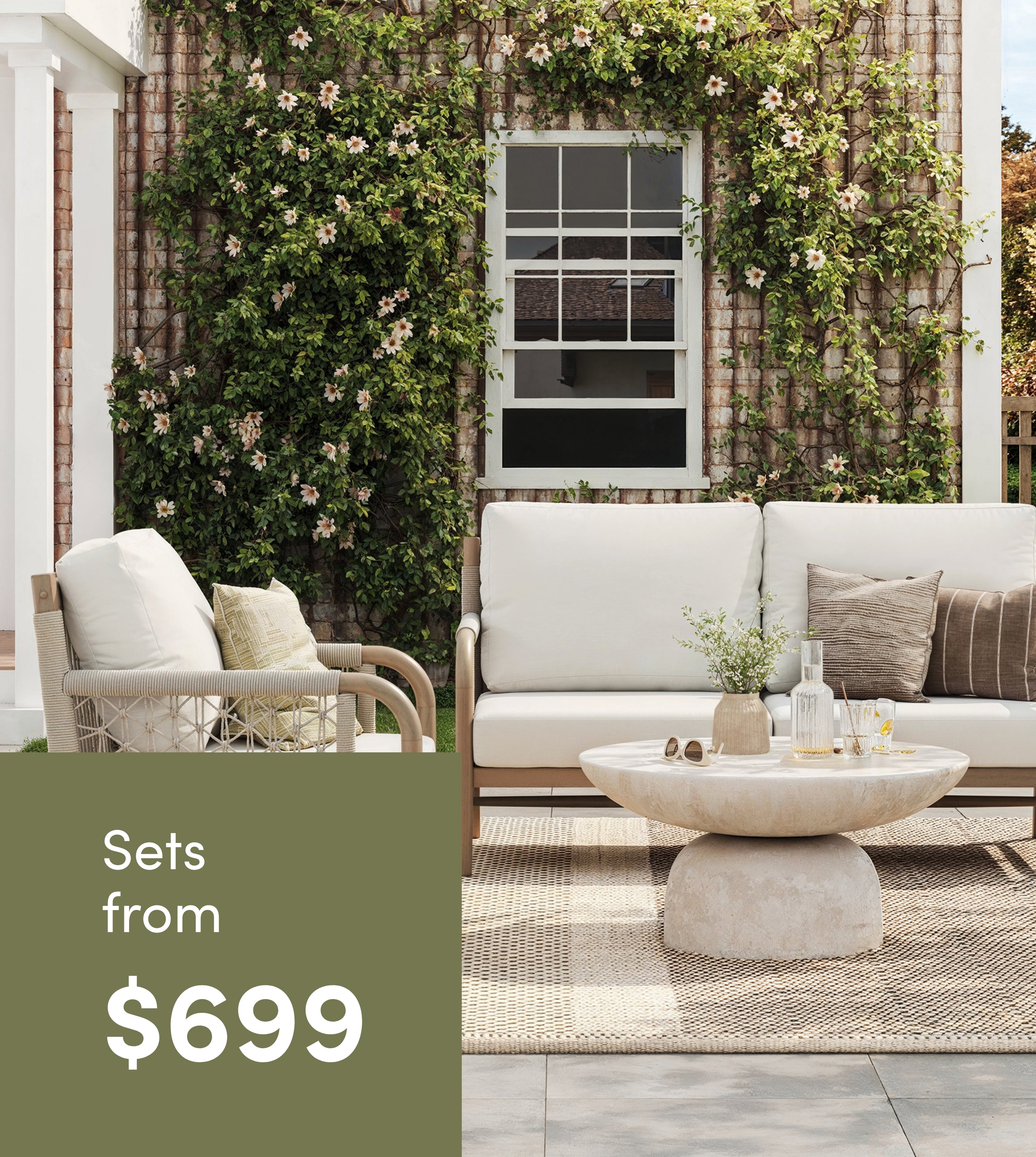 Outdoor patio furniture set with cushioned sofa, armchair, and round coffee table on a rug, in front of a brick wall covered with green vines and flowers. Text reads: Sets from $699.