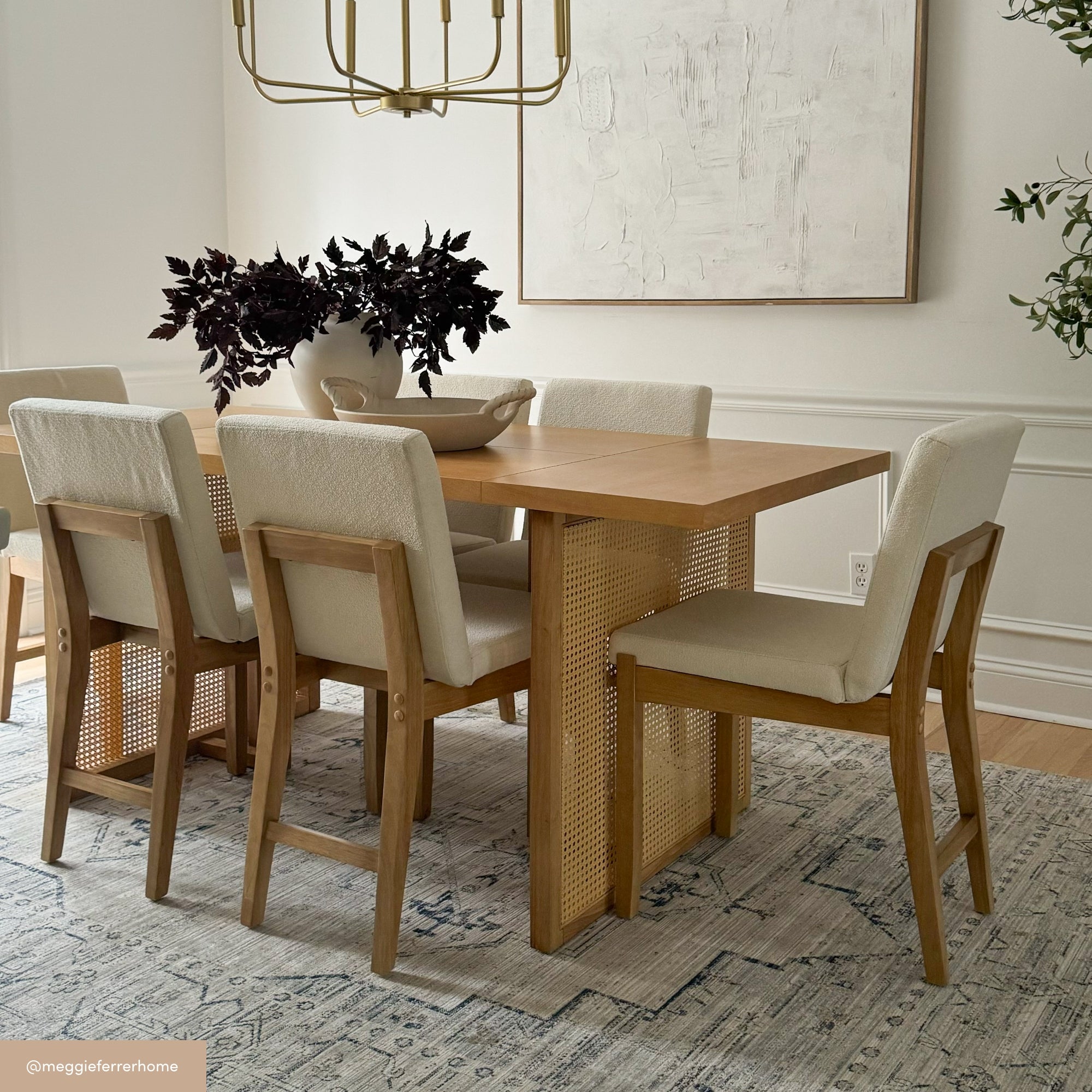 A modern dining room with a wooden table, six light upholstered chairs, a white bowl with dark leafy branches as a centerpiece, a large rug, and a gold chandelier above. Light walls and greenery complete the space.
