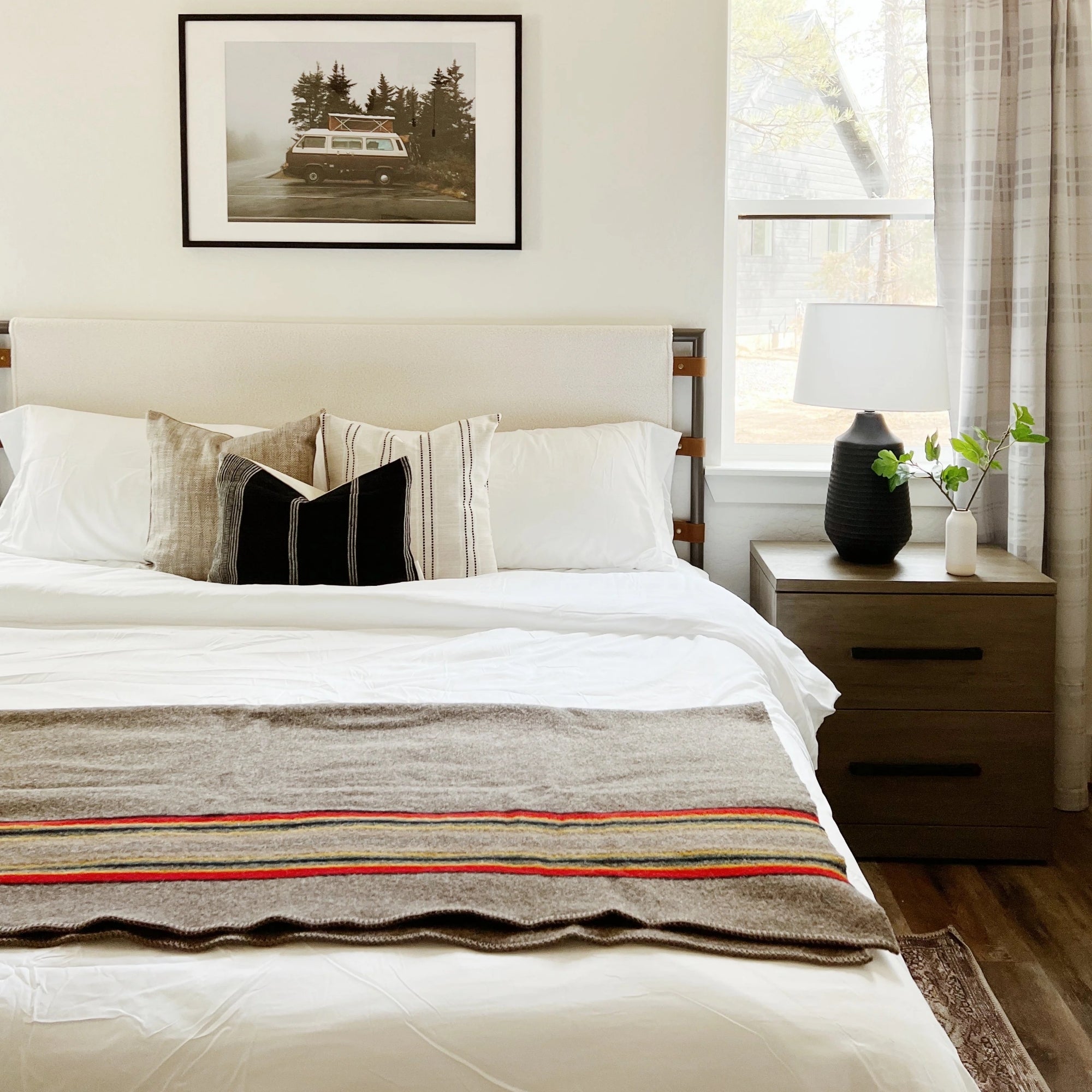 A neatly made bed with white bedding, decorative pillows, and a striped throw blanket sits beside a wooden nightstand with a black vase, a lamp, and leafy branches; a framed photo hangs above the bed.