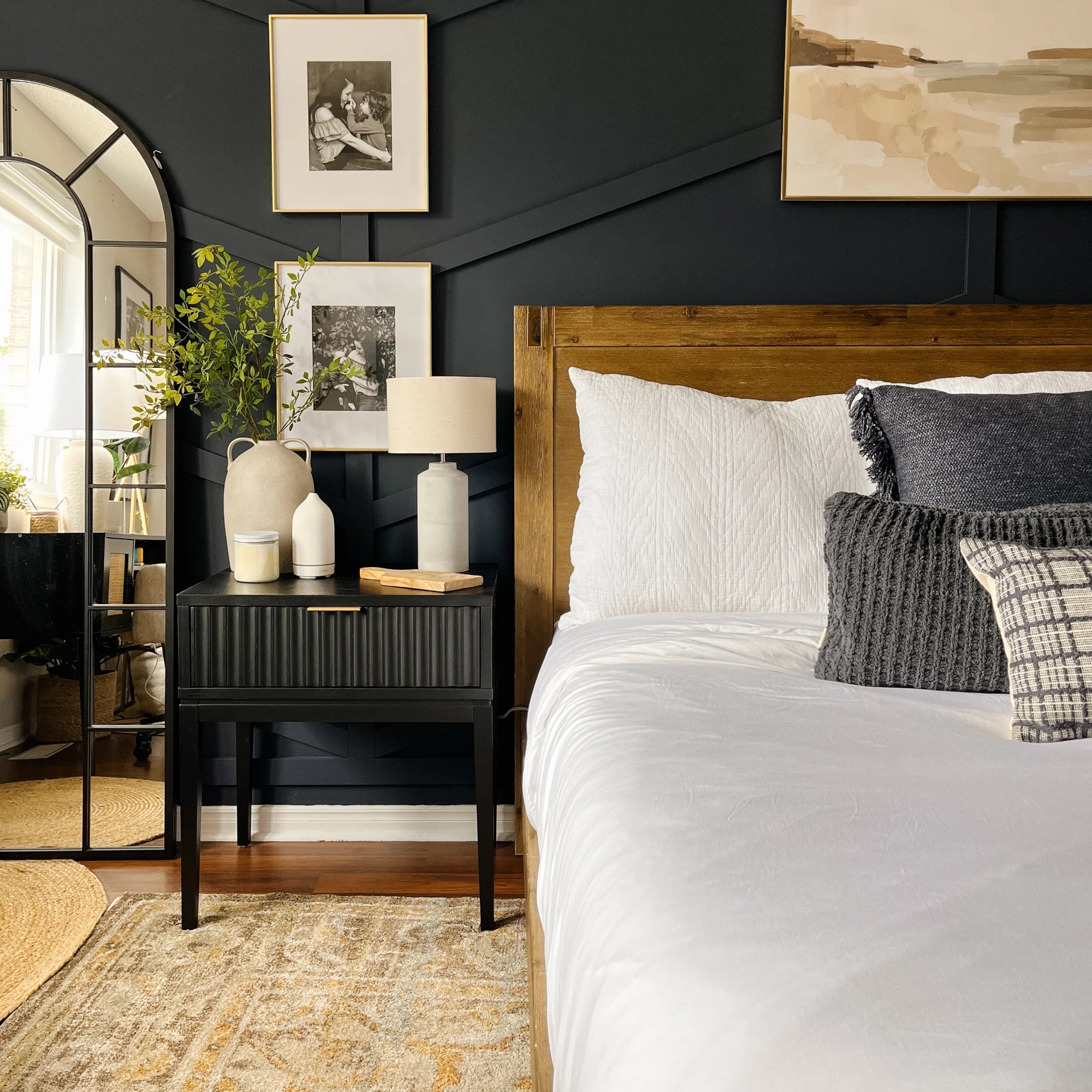 A modern bedroom with a dark accent wall, wooden bed frame, white bedding, and assorted gray pillows. A black nightstand holds a lamp and vases. A tall mirror reflects plants and wall art decor.