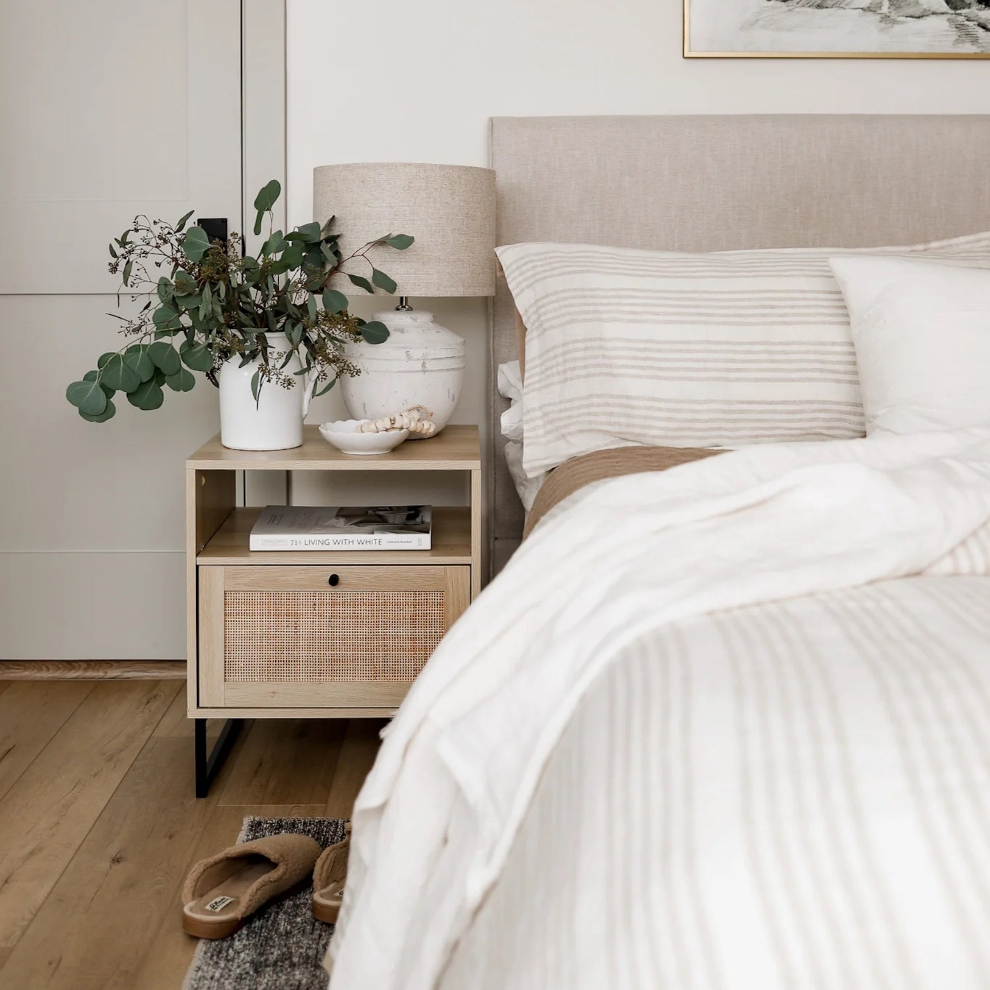 A cozy bedroom scene with a beige upholstered bed, white and beige striped bedding, a wooden nightstand with a drawer, a lamp, a vase with greenery, a bowl, and a book. Slippers are on a rug beside the bed.