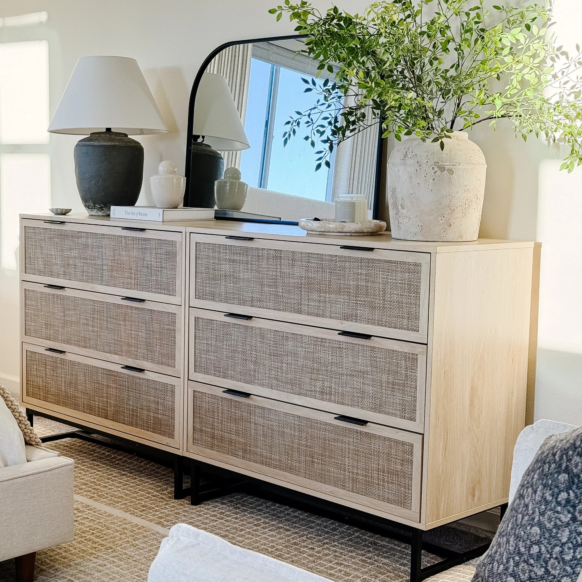 A wooden dresser with woven drawer fronts is topped with a large ceramic lamp, decorative vases, stacked books, a candle, a large mirror, and a tall vase with green branches. The setting is bright and modern.