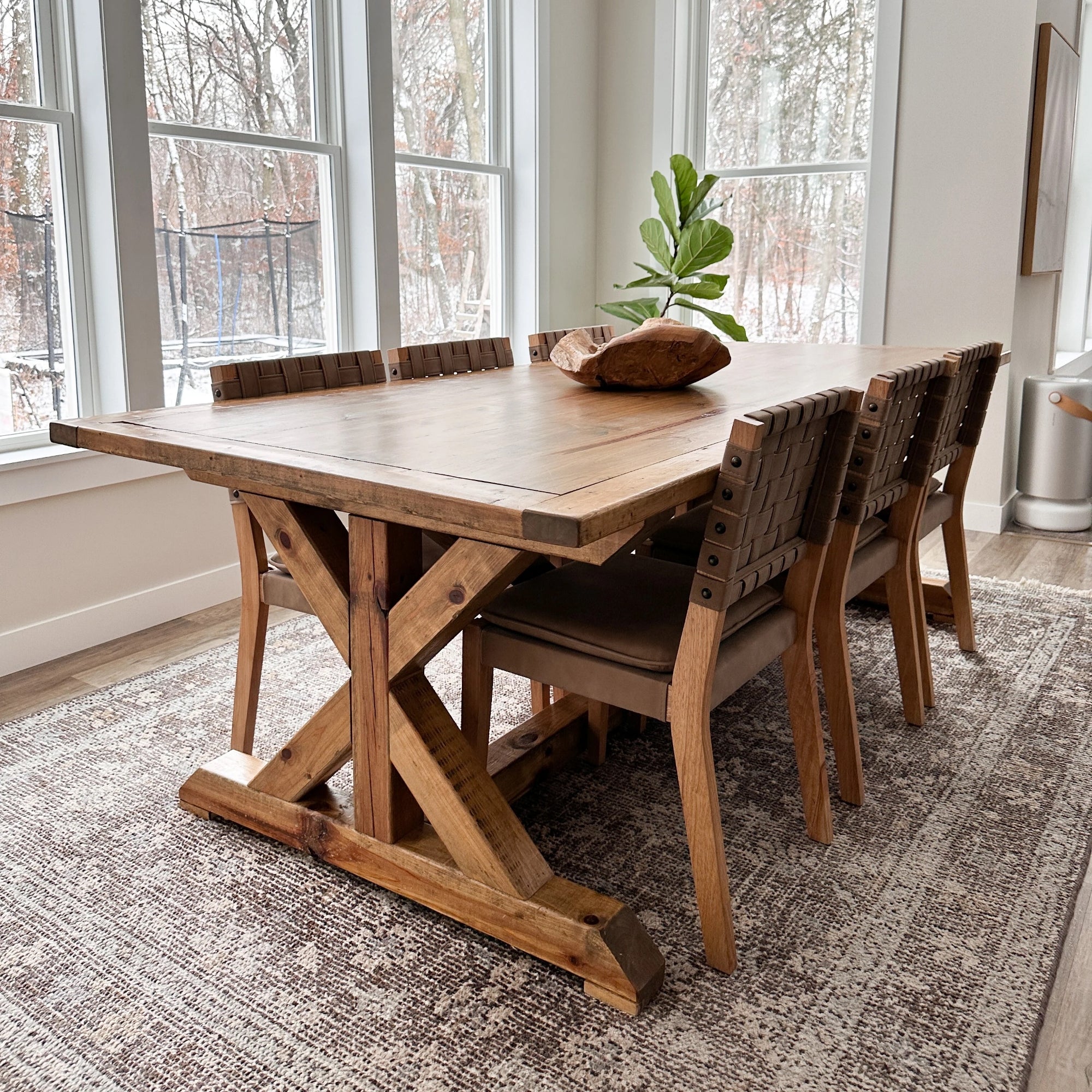 A wooden dining table with a rustic trestle base is surrounded by six woven-back chairs. A decorative wooden bowl and a green plant sit on top. Large windows reveal a snowy outdoor scene. The room features a patterned rug.