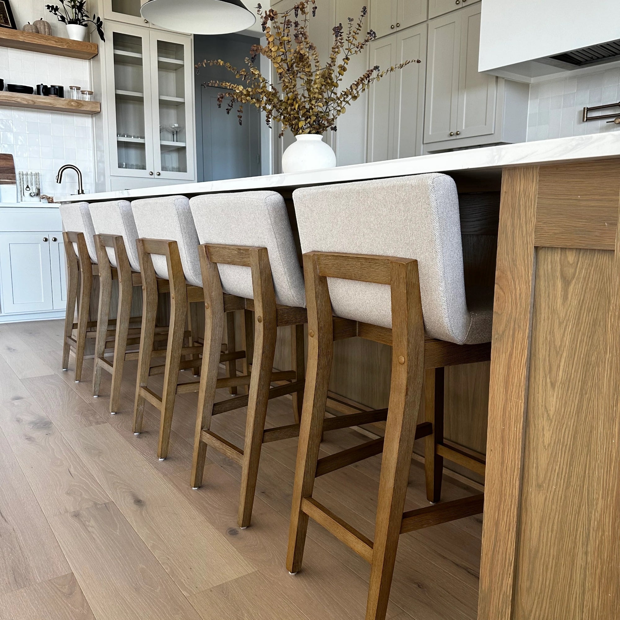 Five wooden bar stools with light upholstered backs are lined up along a kitchen island with a marble countertop. A vase with dried flowers sits on the counter, and white cabinets are visible in the background.