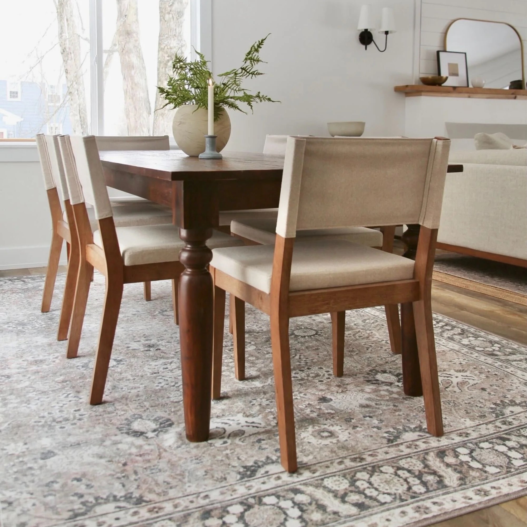 A wooden dining table with four beige upholstered chairs sits on a patterned rug in a bright dining room with large windows, a vase with greenery, and a candle as centerpiece. A neutral living area is visible in the background.