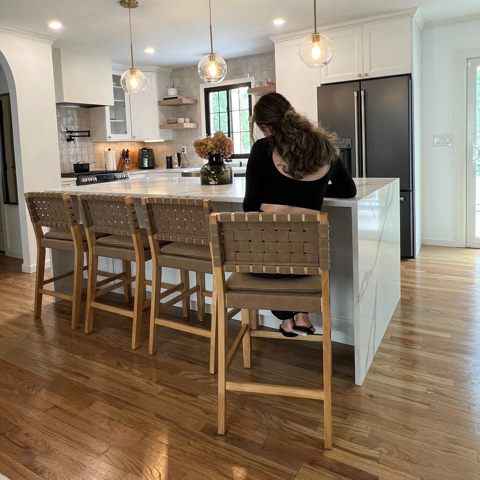 A woman with long hair, wearing a black top, sits alone on a stool at a modern kitchen island with four wooden chairs. The kitchen has white cabinets, pendant lights, and a vase of flowers on the counter.