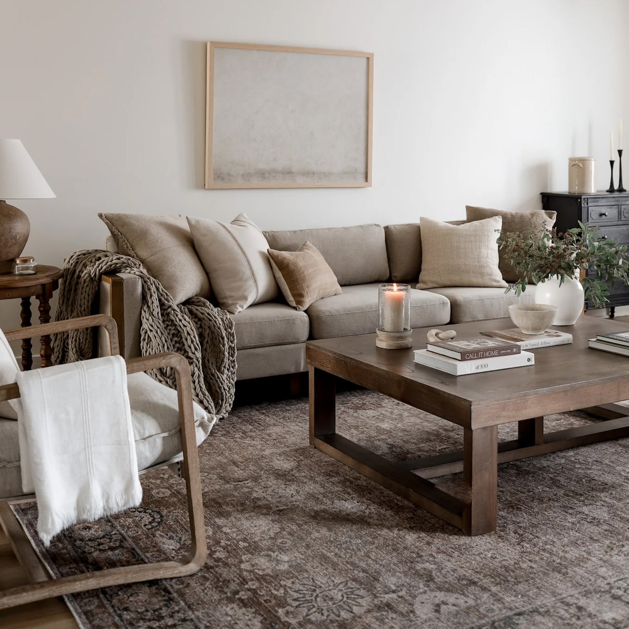 A cozy living room with a beige sectional sofa, neutral pillows, a knitted throw, wooden coffee table with books and a candle, a modern armchair, a table lamp, and minimalist decor in soft, earthy tones.