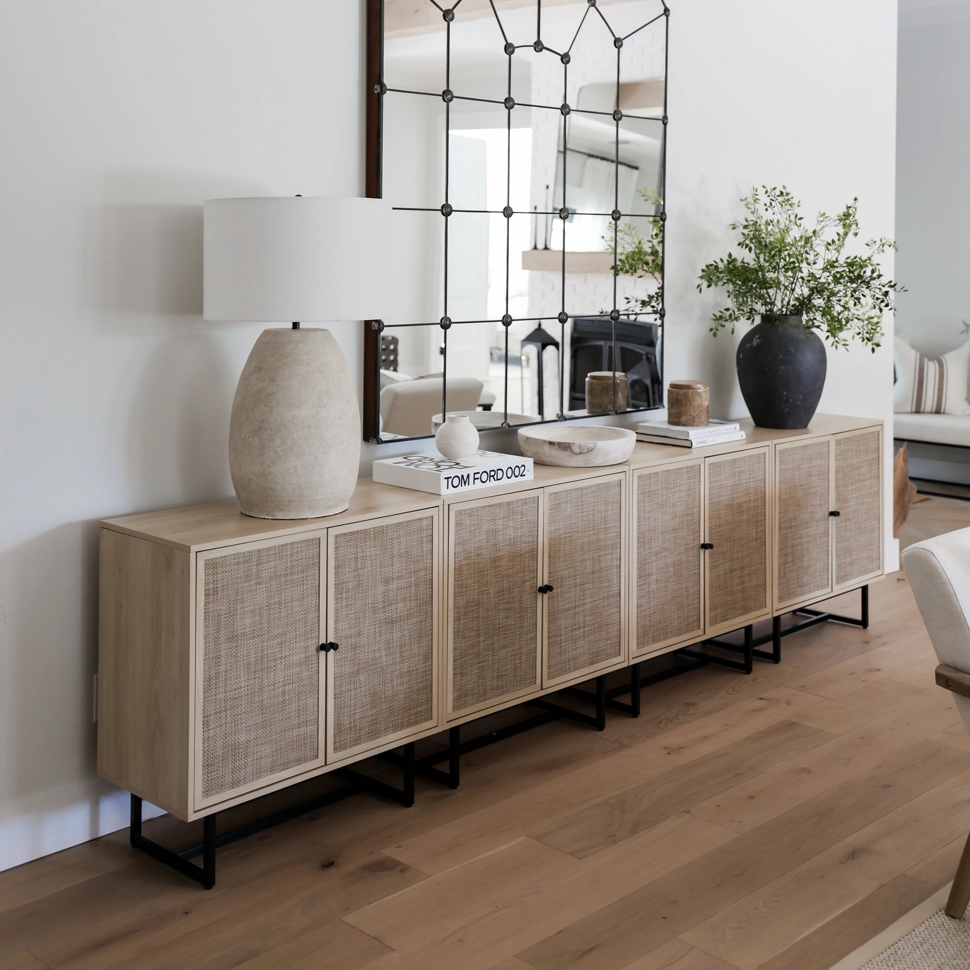 A light wood sideboard with rattan panels stands against a white wall, topped with a large mirror, a stone lamp, a black vase with greenery, and decorative objects, in a modern, minimalist living space.
