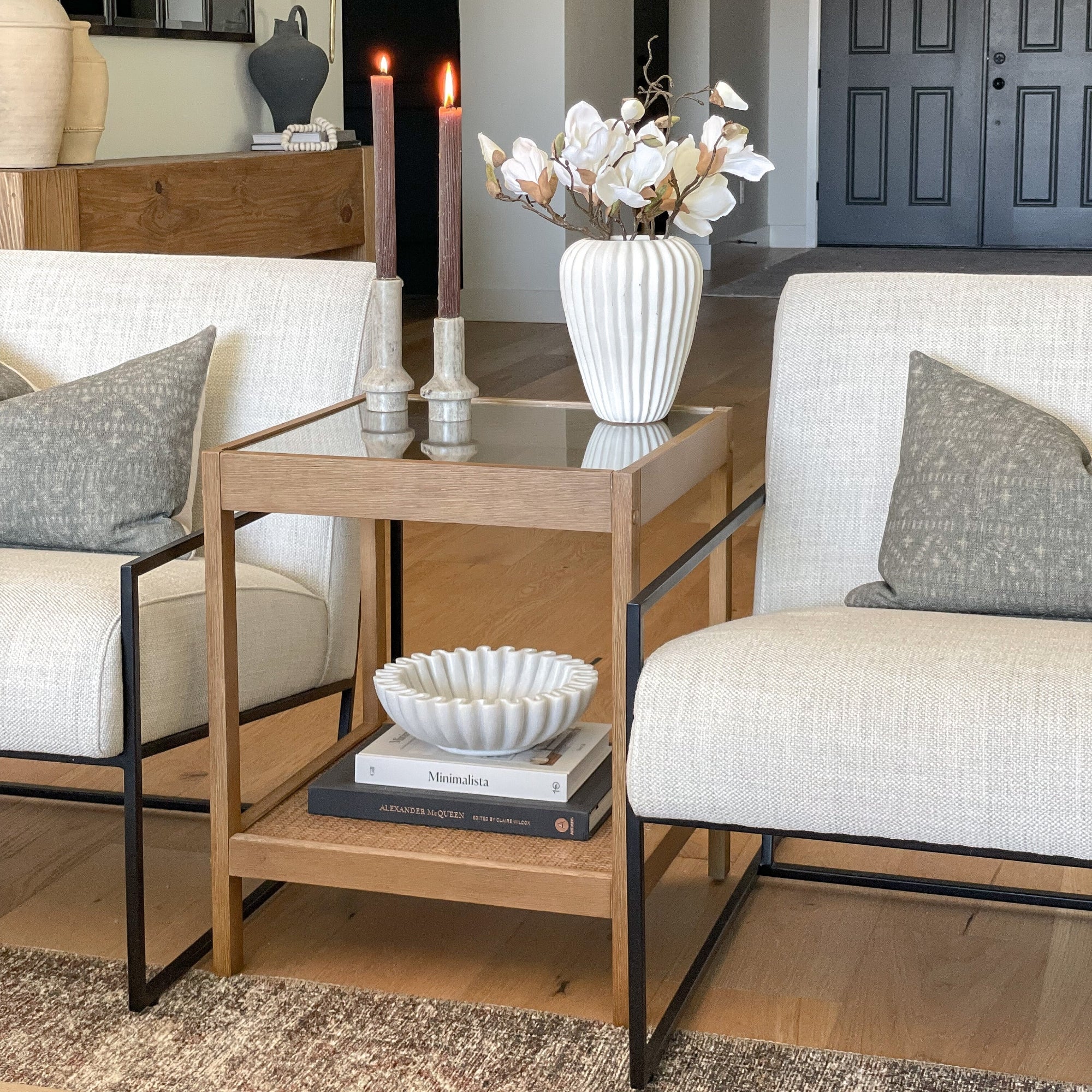 A modern living room scene with two beige chairs, a wooden side table holding a white vase with flowers, two candles, and decorative books. A dark bowl and books are displayed on the lower shelf of the table.
