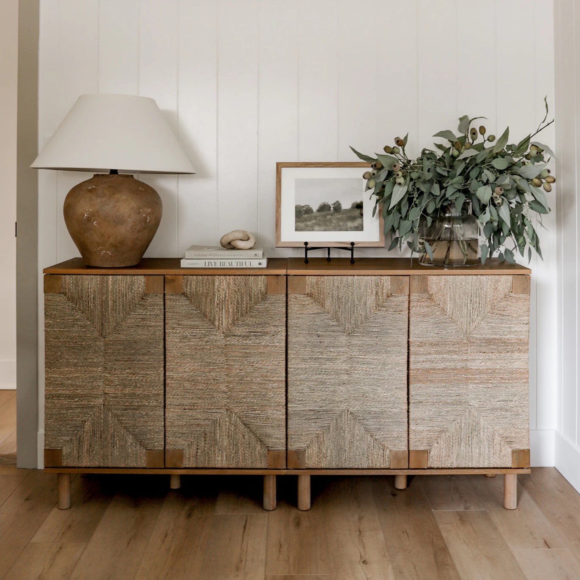A woven textured sideboard holds a brown lamp with a white shade, a stack of books, a framed photo, and a vase filled with leafy greenery, set against a white paneled wall on a wood floor.