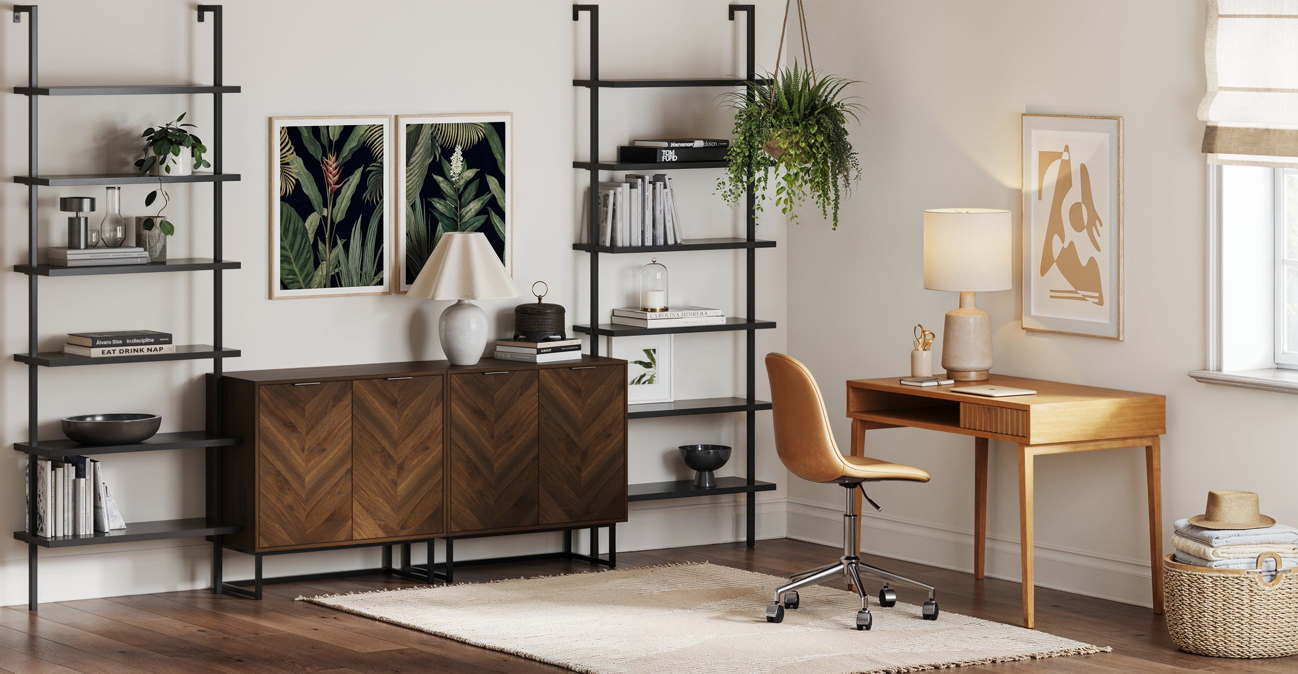 A modern home office with wood furniture, open black shelves with books and decor, a desk with a lamp, an office chair, framed art on the wall, a hanging plant, and a woven rug on hardwood floors.
