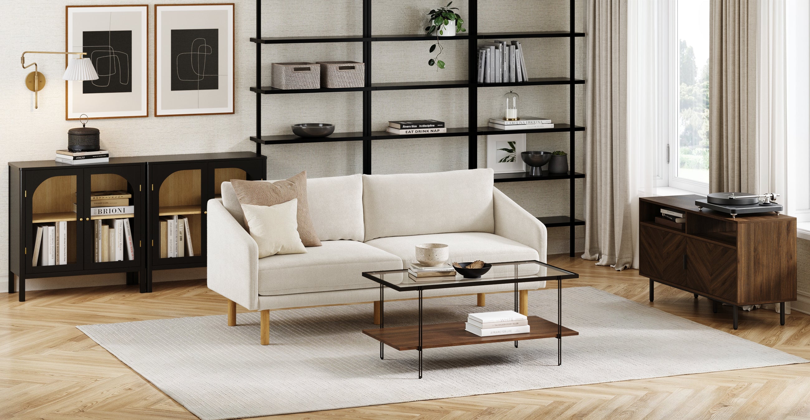 Modern living room with a light beige sofa, glass coffee table, large black shelving unit, cabinets with books, minimalist wall art, and a turntable cabinet by a window with beige curtains. Light wood herringbone floor and neutral rug.