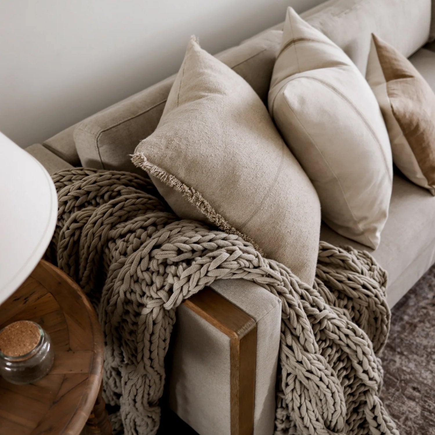 A cozy beige sofa with three large, textured pillows and a chunky knit blanket draped over the armrest, next to a round wooden side table with a small jar and a lamp.