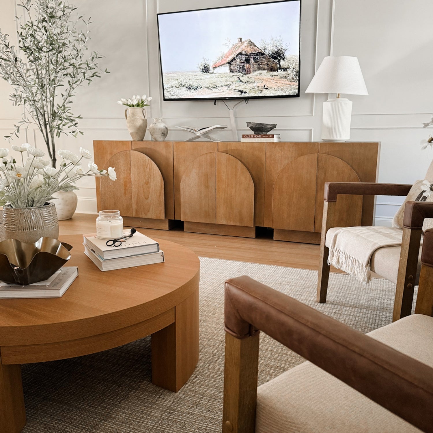 A modern living room with a wooden coffee table and sideboard, neutral armchairs, a TV displaying a cottage scene, decorative vases, books, and plants, all in warm, beige and wood tones.
