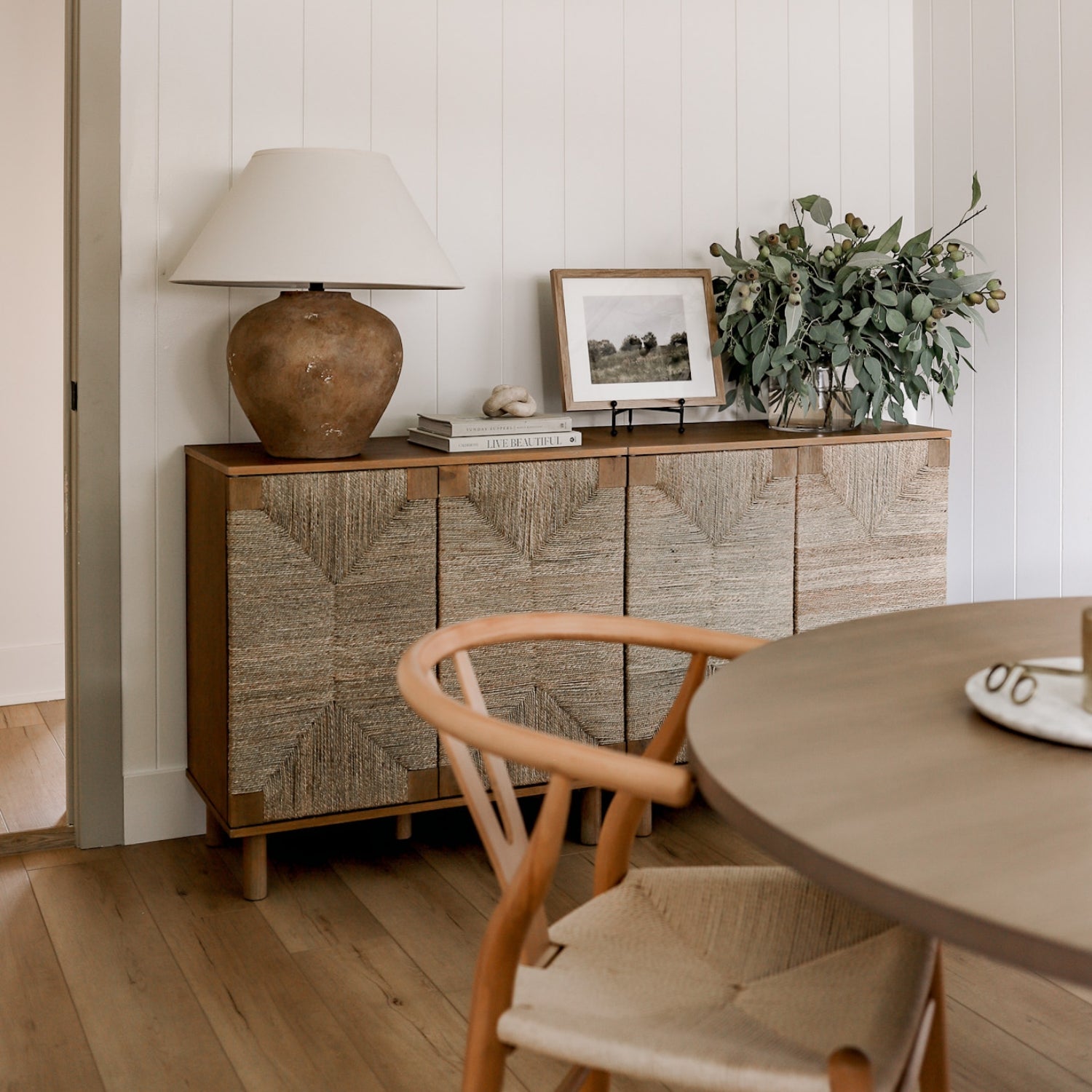 A cozy dining space with a round wooden table, woven chair, and a sideboard decorated with a large ceramic lamp, framed photo, books, and a vase of leafy branches against white paneled walls and wood flooring.