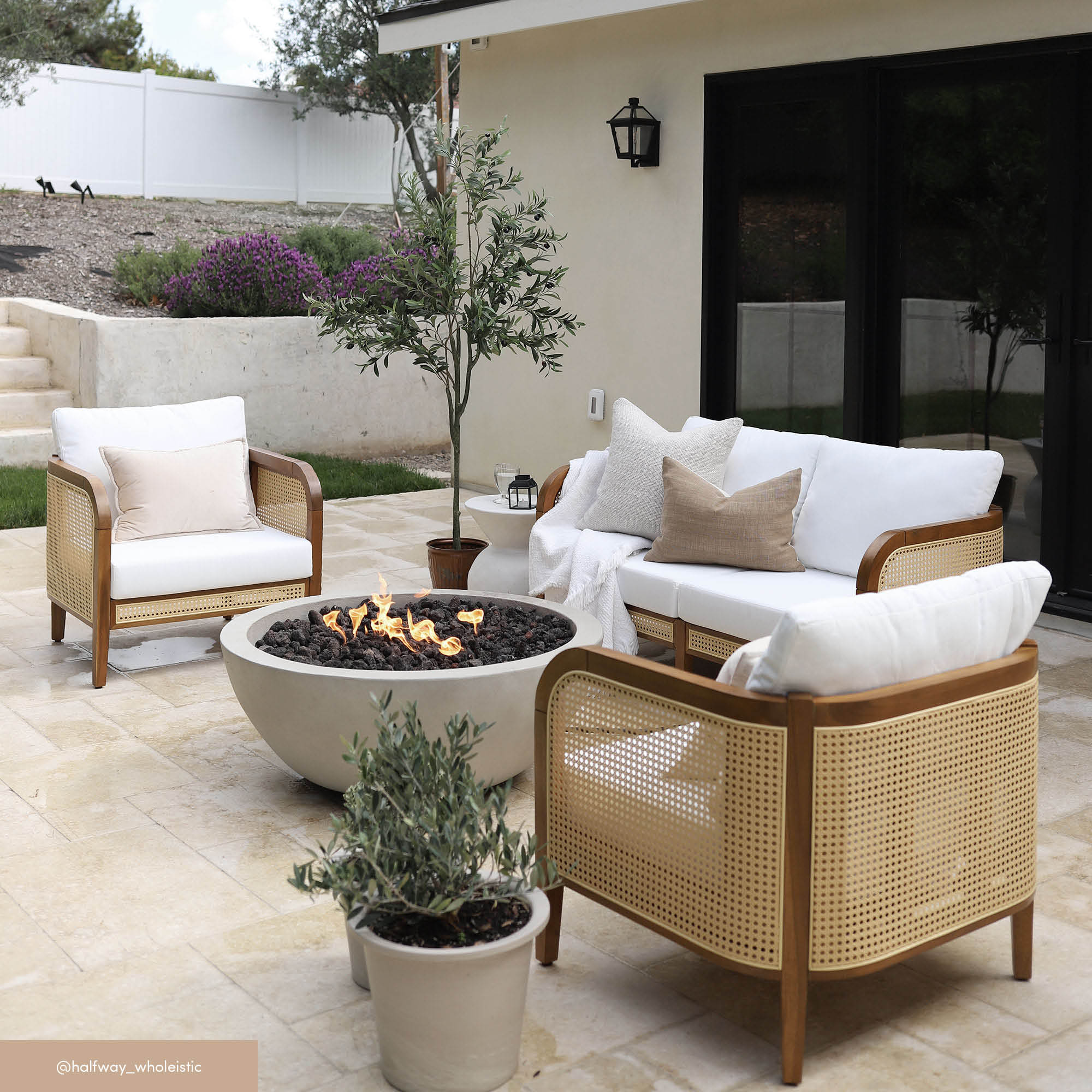 Modern outdoor patio with a cream sofa, two armchairs with cane sides, neutral cushions, and a round concrete fire pit. Potted plants and a small tree add greenery to the light stone flooring.