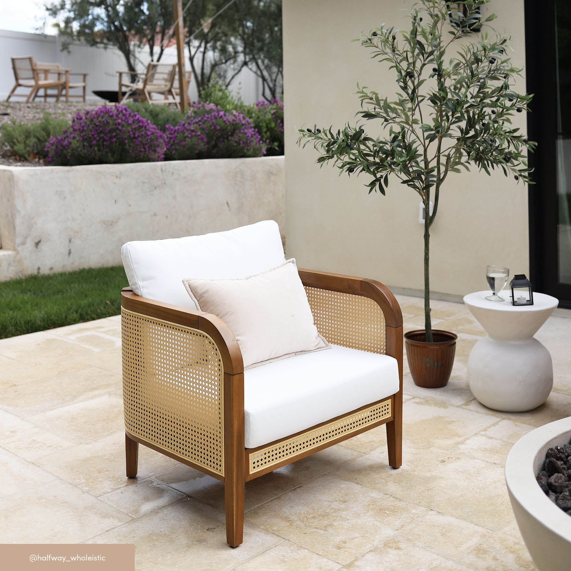 A patio with a cushioned rattan armchair, a small round table holding two glasses and a book, a potted tree, and a landscaped garden with more seating in the background.