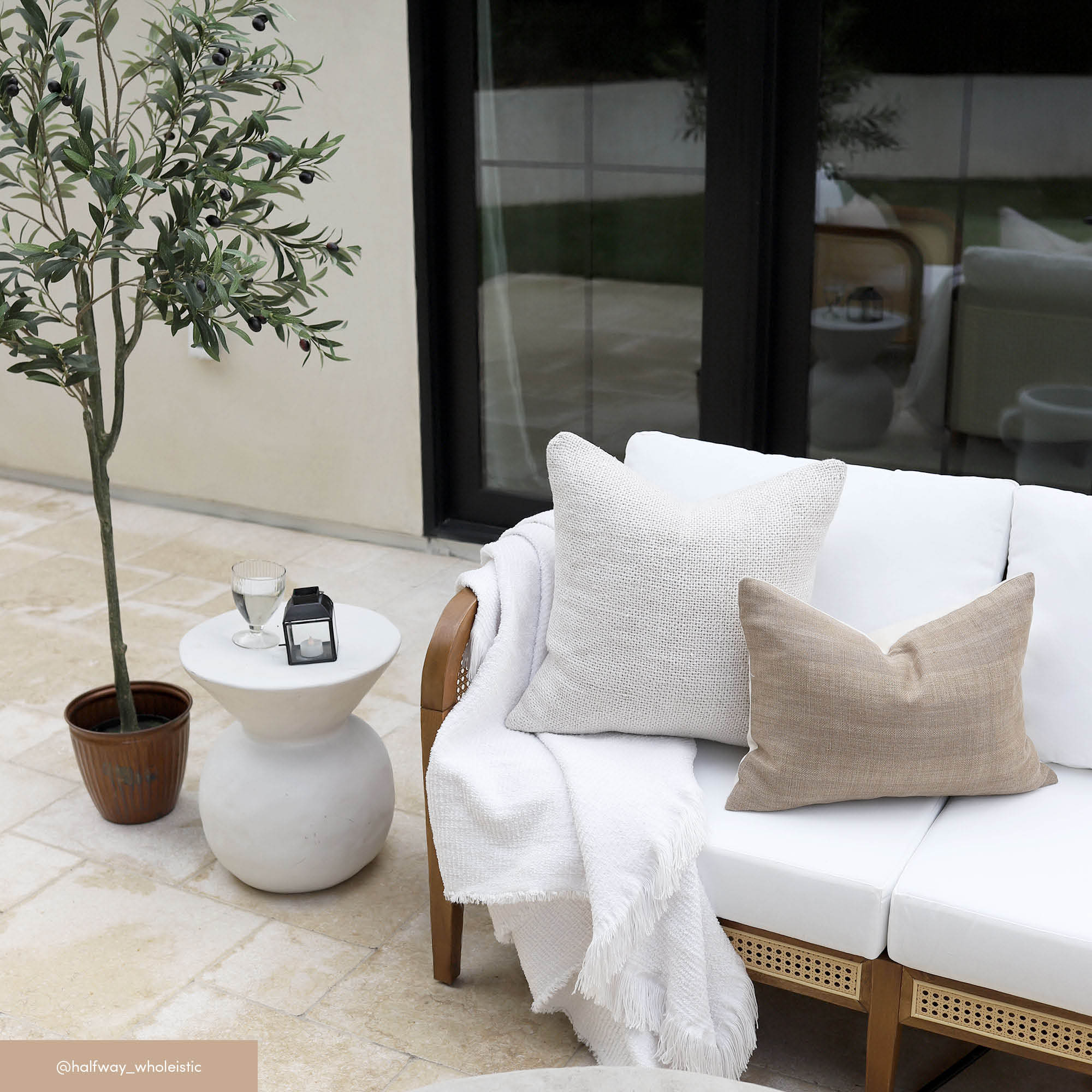 A modern outdoor patio with a white cushioned sofa, beige and white pillows, and a white blanket. A small round table holds a glass, a candle, and a potted olive tree stands nearby. Large windows reflect the scene.