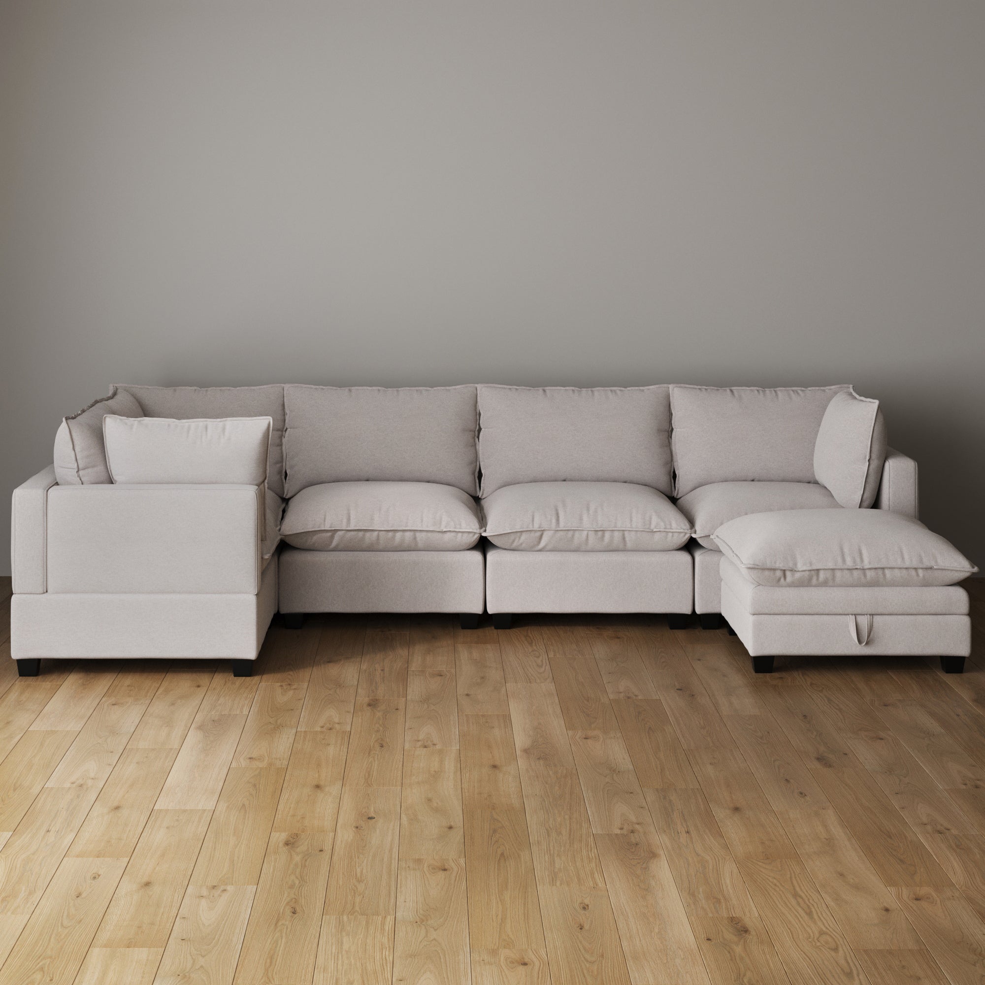 A light beige sectional sofa with plush cushions is arranged against a plain gray wall on a light wood floor in a minimalist room.