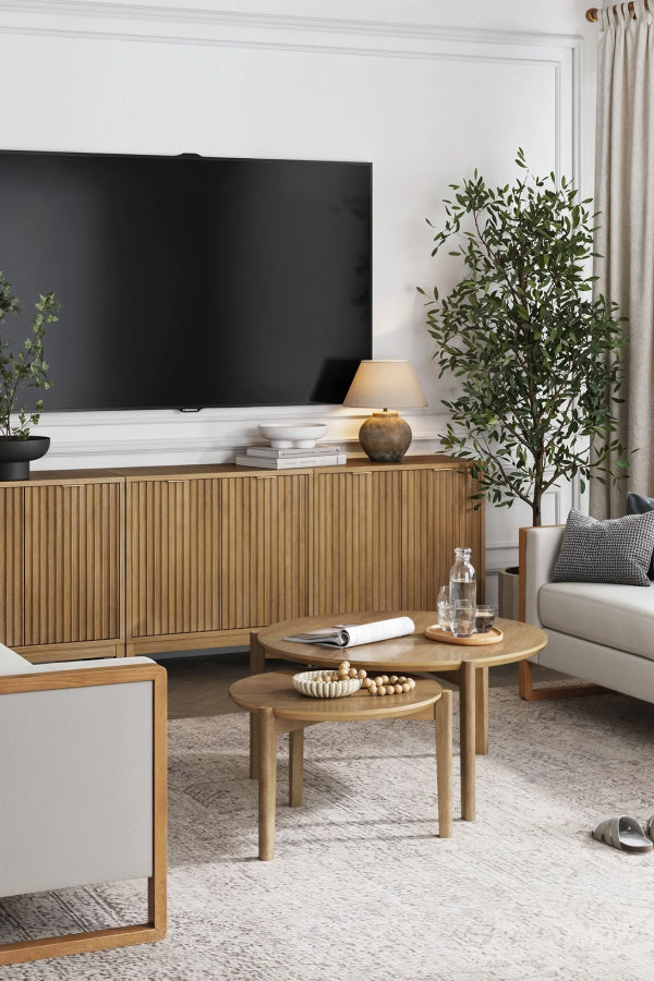 A modern living room with a large flat-screen TV, a wooden console, two round wooden coffee tables, a cream sofa, potted plants, a table lamp, and neutral decor in soft, natural tones.