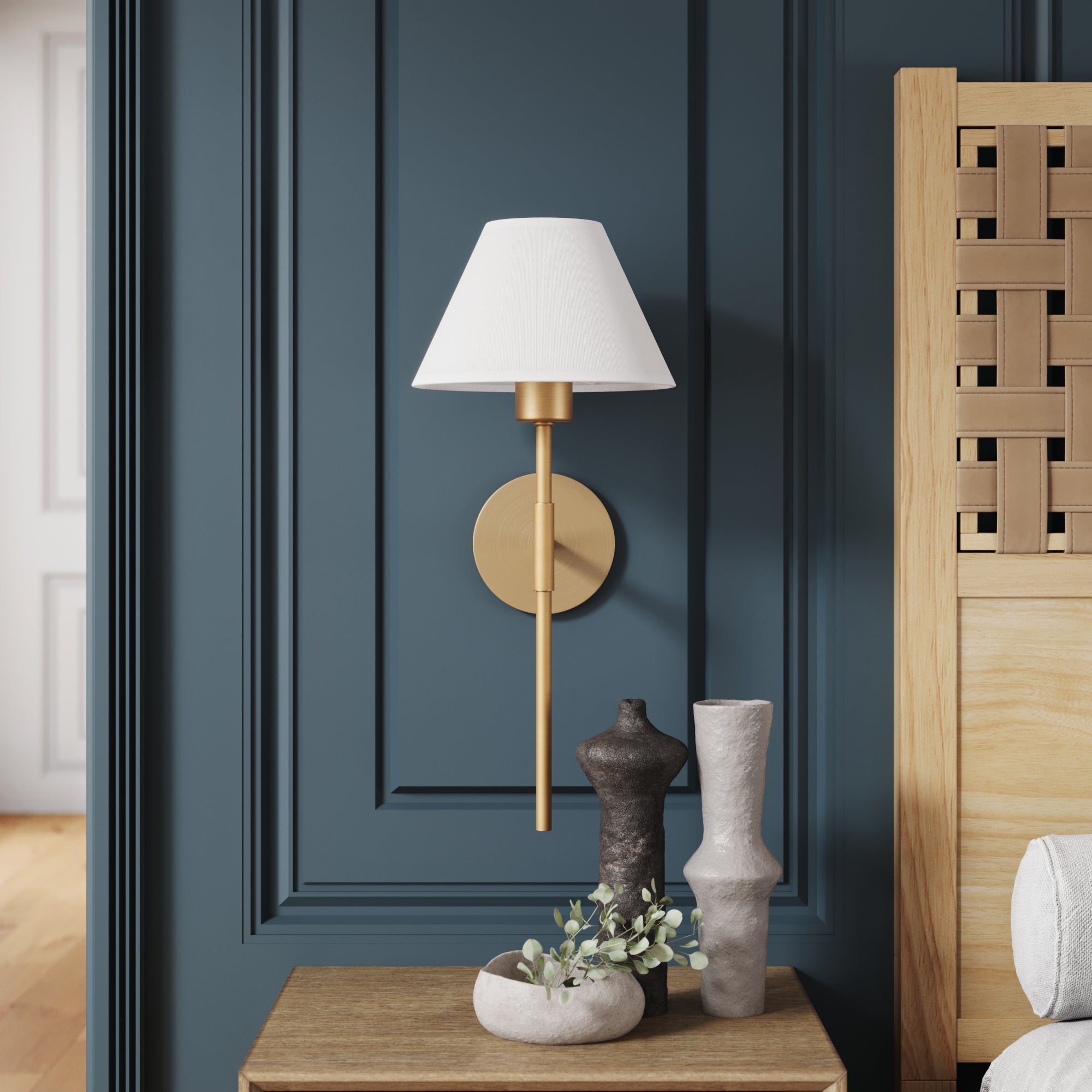A gold wall sconce with a white lampshade is mounted on a dark blue paneled wall above a wooden nightstand, which holds abstract vases and a small plant.
