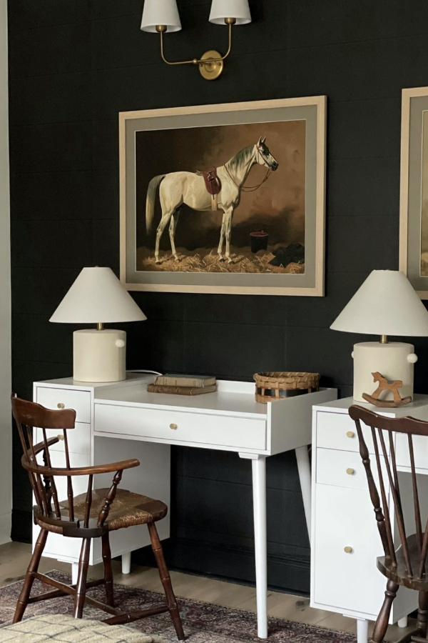 A white desk with matching lamps sits against a dark wall, topped with books and a small basket. Above the desk hangs a framed painting of a white horse with a saddle. Two wooden chairs are positioned in front of the desk.