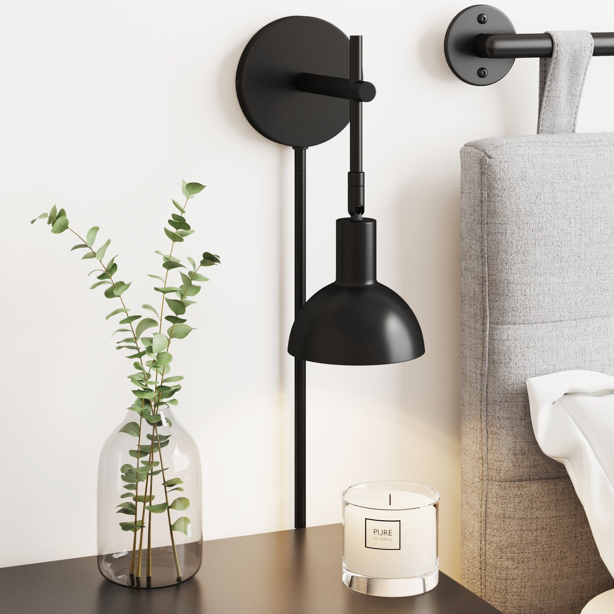 The Nathan James Plug-In Adjustable Metal Wall Light in Black is mounted above a dark nightstand with a clear vase and PURE candle beside a light gray bed—perfect as a renter-friendly wall sconce.