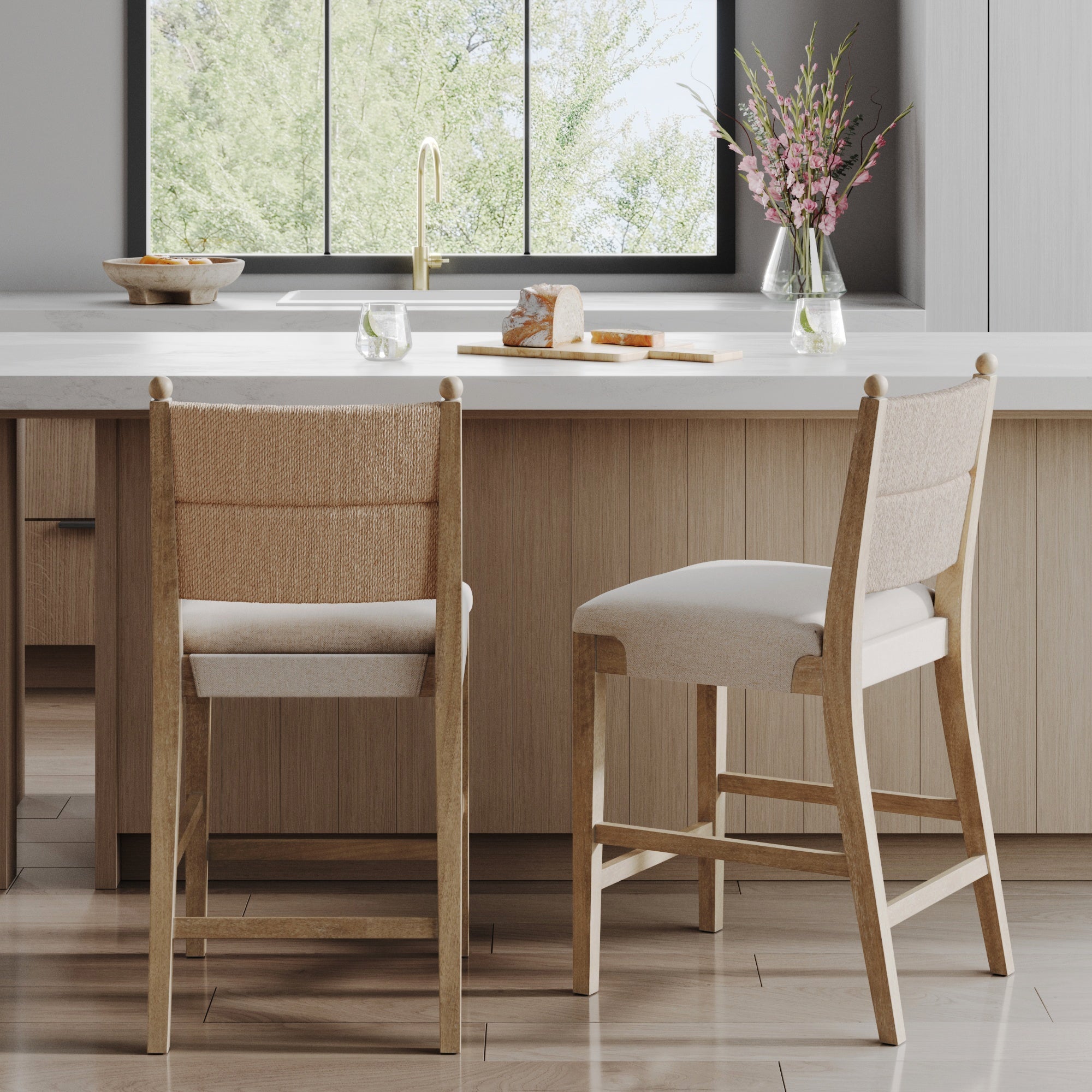 Two Nathan James Seagrass & Wood Counter Height Bar Stools in light brown (set of 2) sit at a modern kitchen island with a vase of pink flowers, bread, and two glasses of water. A large window reveals lush greenery outside.