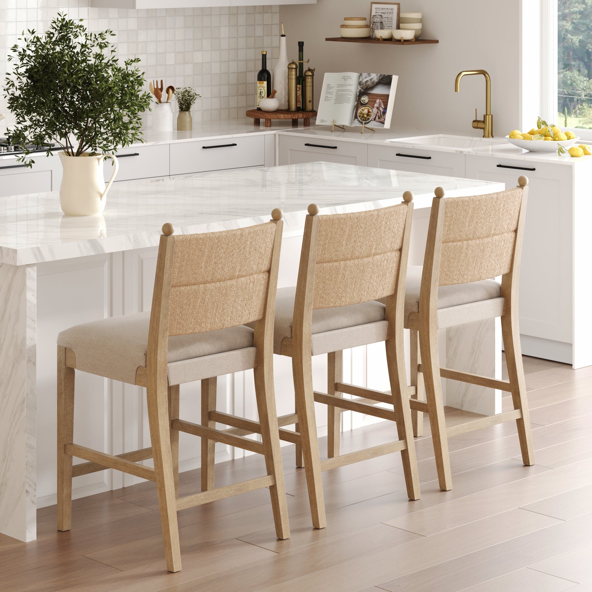 Three Nathan James Seagrass & Wood Counter Height Bar Stools in light brown (set of 3) are placed by a white marble kitchen island in a bright, modern kitchen with plants, books, and kitchenware on the shelves and countertops.