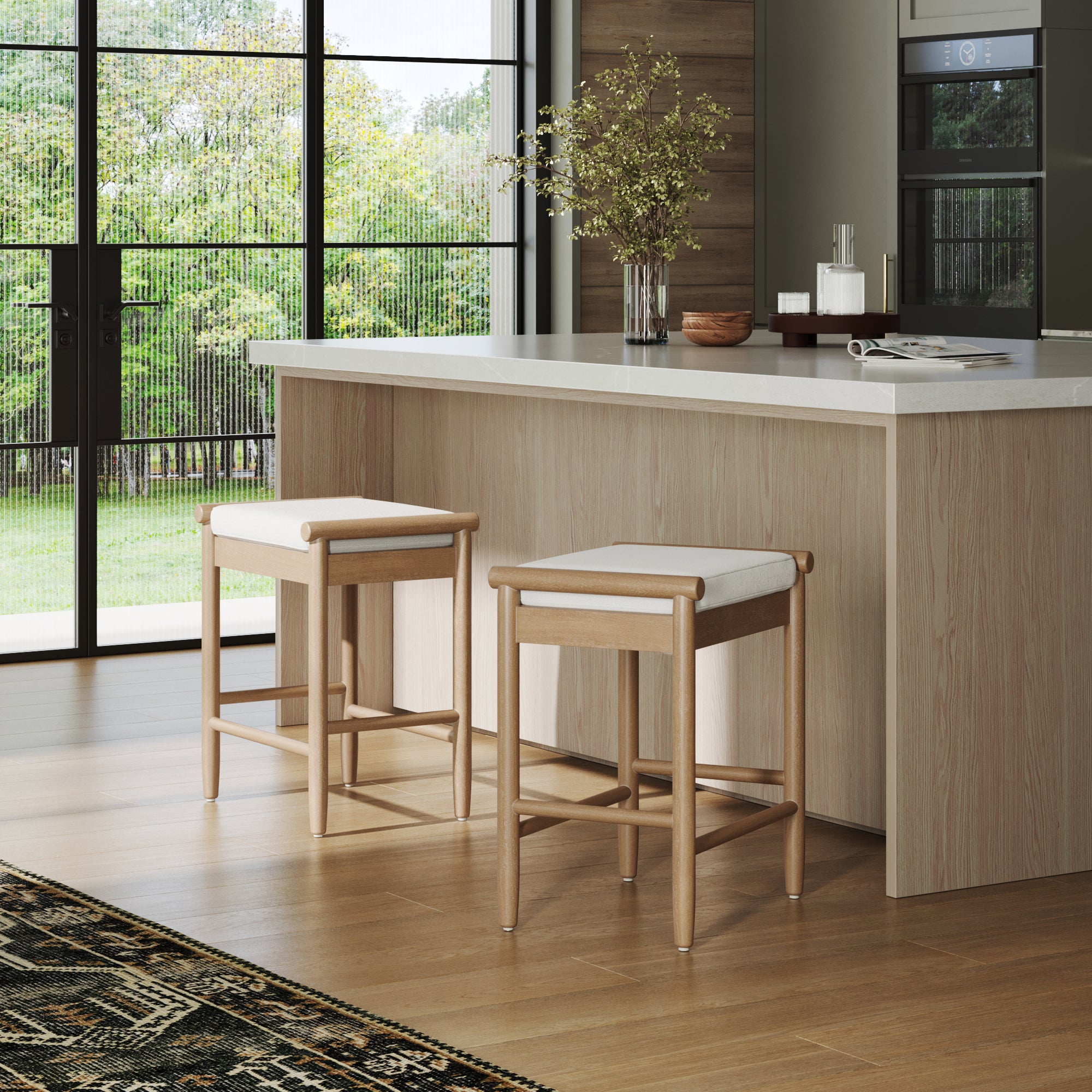 A modern kitchen with light wood floors, a matching island with a white countertop, two Nathan James Backless Wooden Bar Stools with light brown boucle upholstery, a vase of branches, and large windows overlooking a green yard.