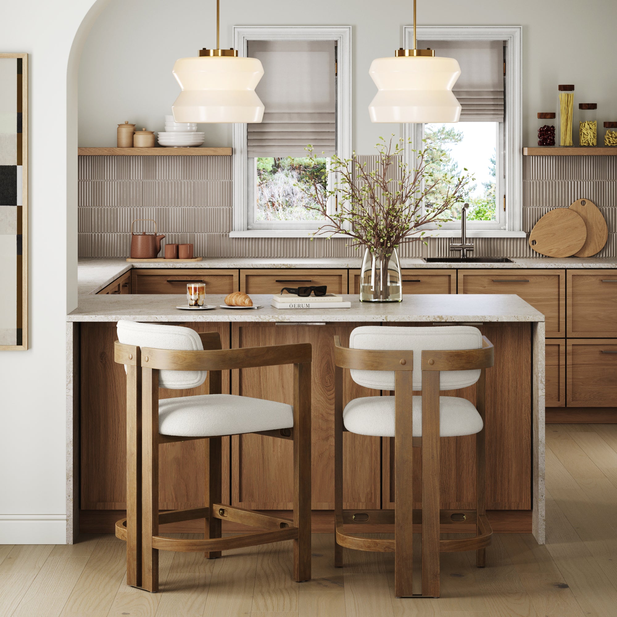Modern kitchen with light wood cabinets, white countertops, and a set of two Nathan James Barrel Bar Stools in light brown at the island. Pendant lights, a vase with branches, breakfast items, and two windows above the sink add charm and natural light.