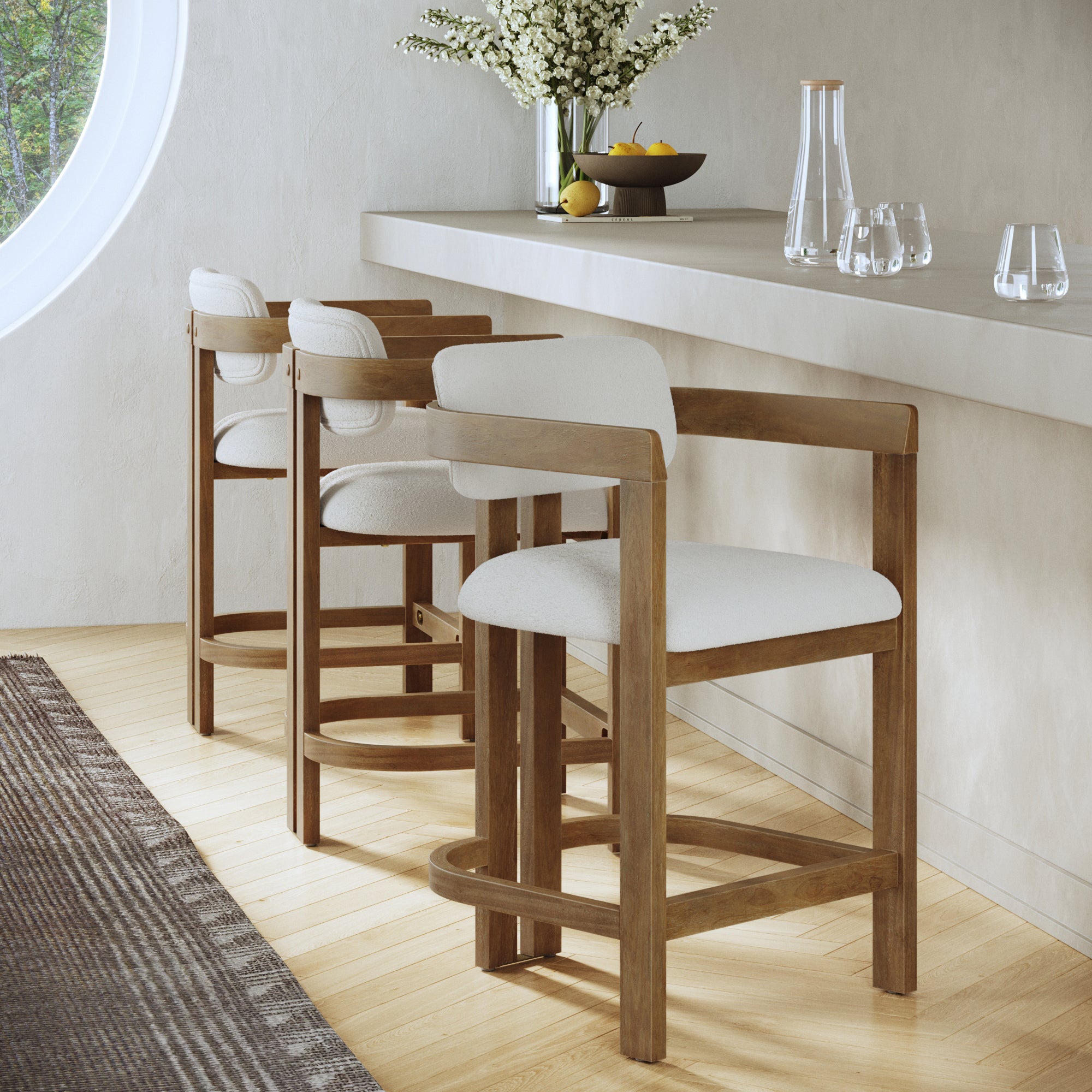 Three Nathan James Barrel Bar Stools with backs and arms in light brown line a pale counter. A vase of white flowers, fruit bowl, and glassware sit atop as sunlight streams through a round window for a bright, modern vibe.