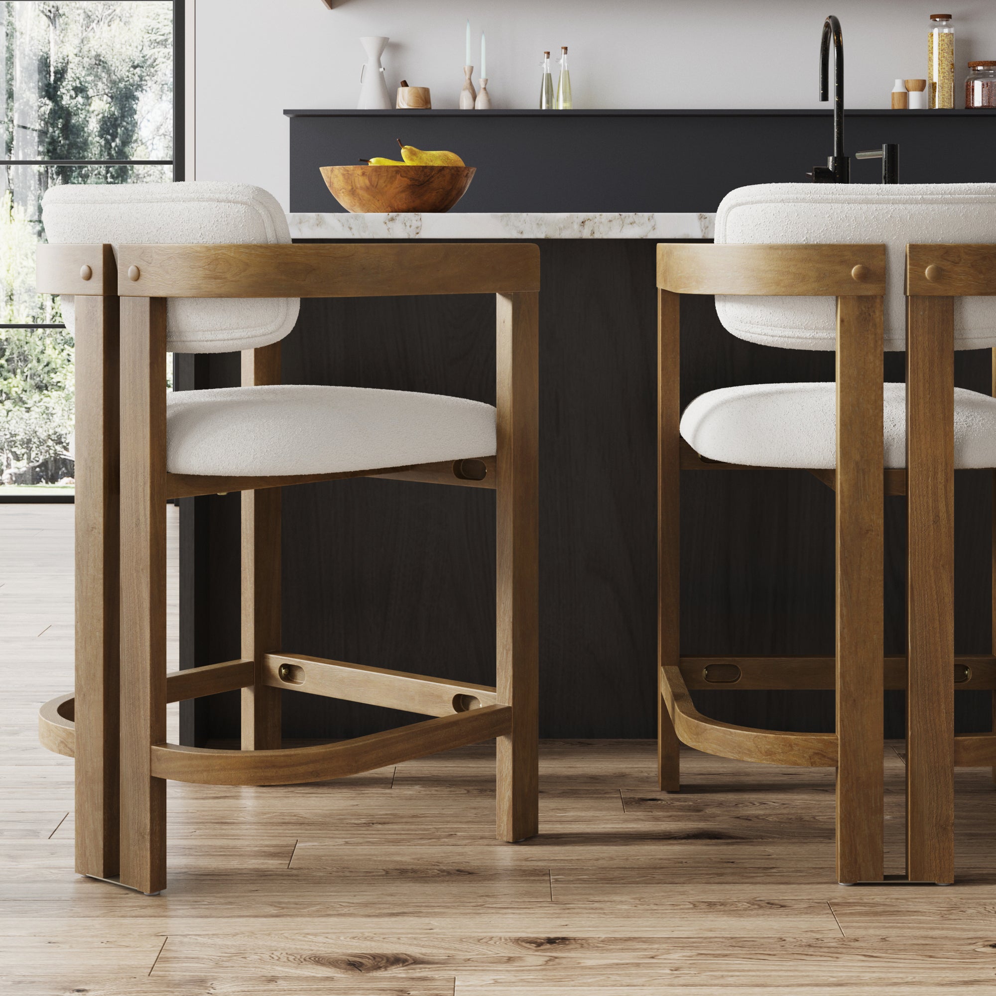 Two Nathan James Barrel Bar Stools w/ Back and Arm in light brown wood and white upholstery are placed at a dark kitchen island with a marble countertop. A wooden fruit bowl and assorted items sit on the counter.