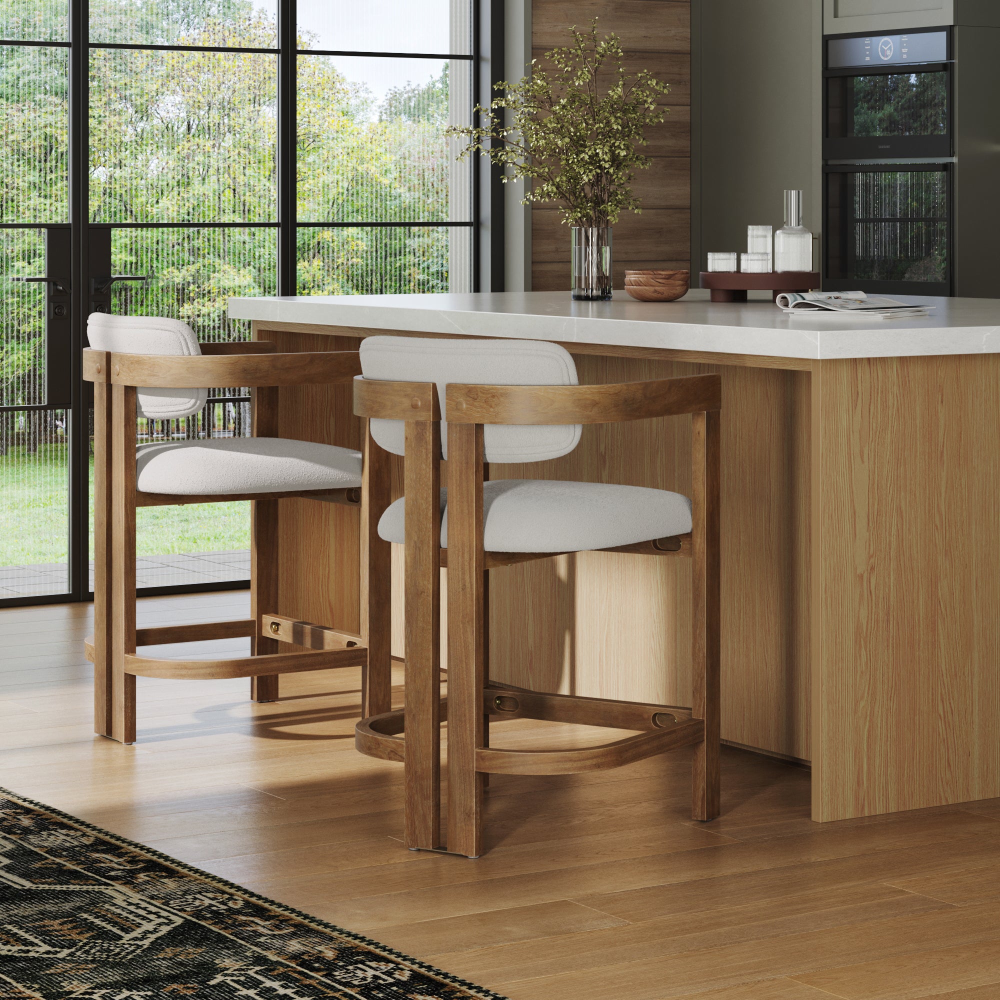 Two Nathan James Barrel Bar Stools with backs and arms in light brown are placed at a light wood kitchen island in a modern space with large windows, indoor plants, and a patterned black rug.