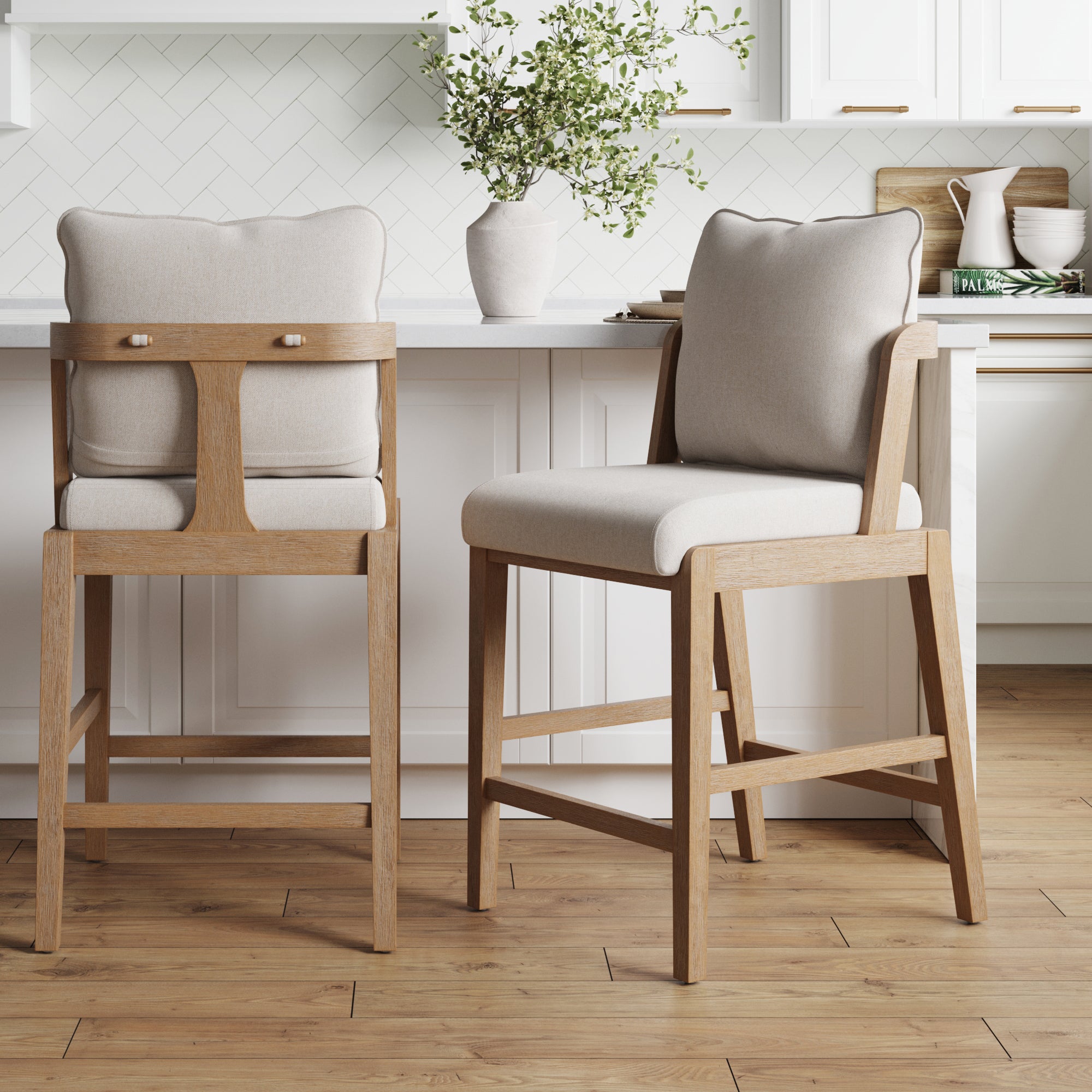 Two Nathan James Scandinavian 2-Pack Bar Stools with Back in light brown, featuring solid wood frames and beige upholstered seats, stand in a modern kitchen with white cabinets, white tile backsplash, and light wood floors.