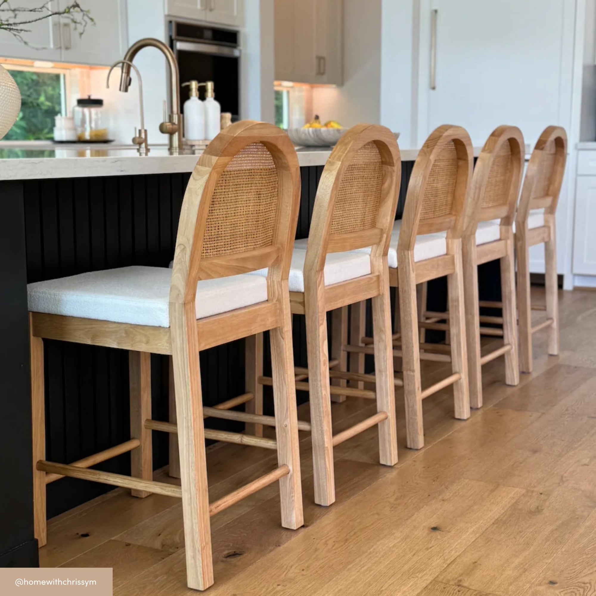 Four wooden bar stools with curved, woven cane backs and white cushions are lined up at a kitchen island with a black base, in a modern kitchen with light wood flooring and white cabinetry.