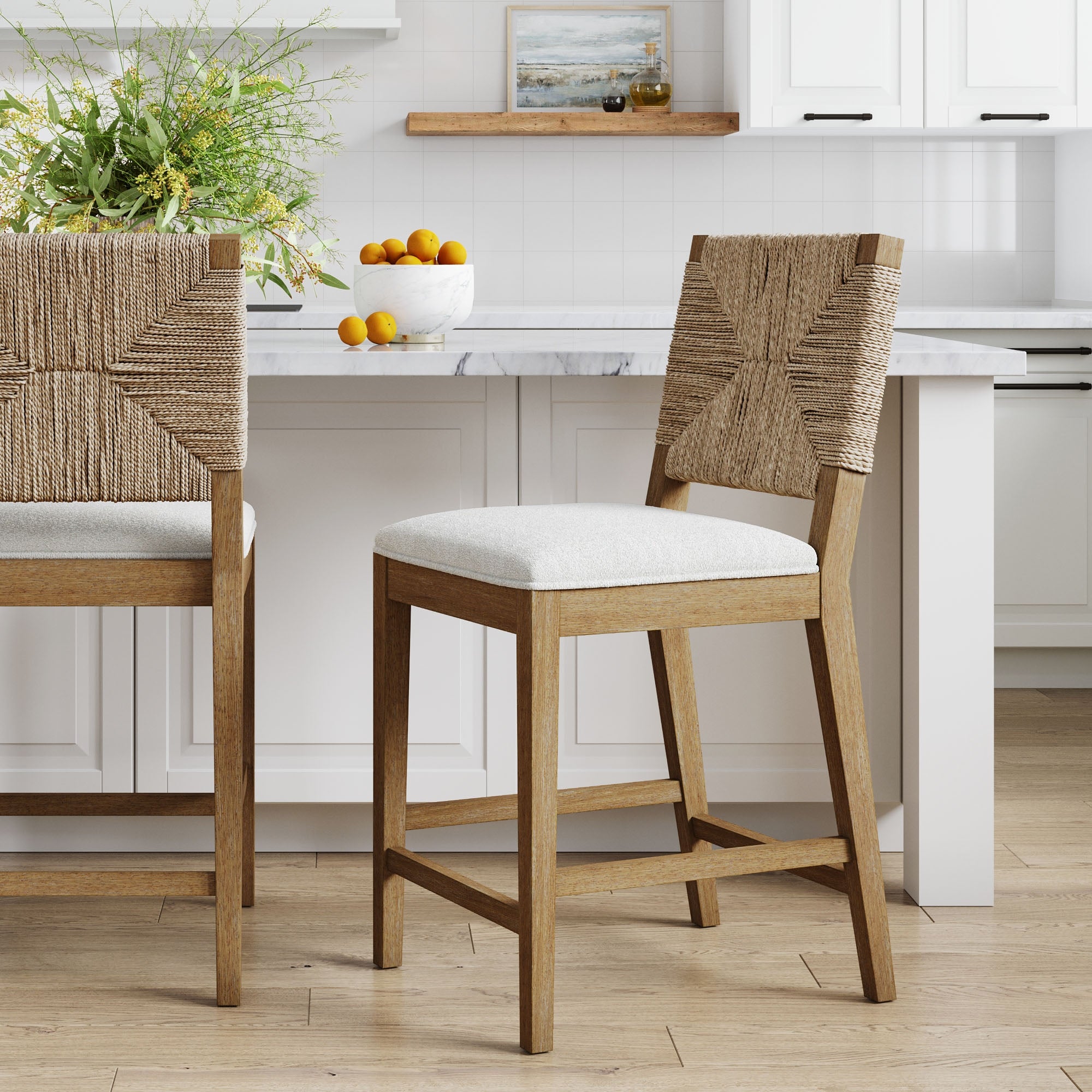 Two wooden counter-height chairs with woven rope backs and cushioned seats are placed in a bright, modern kitchen with a white island, bowl of oranges, greenery, and white cabinetry in the background.