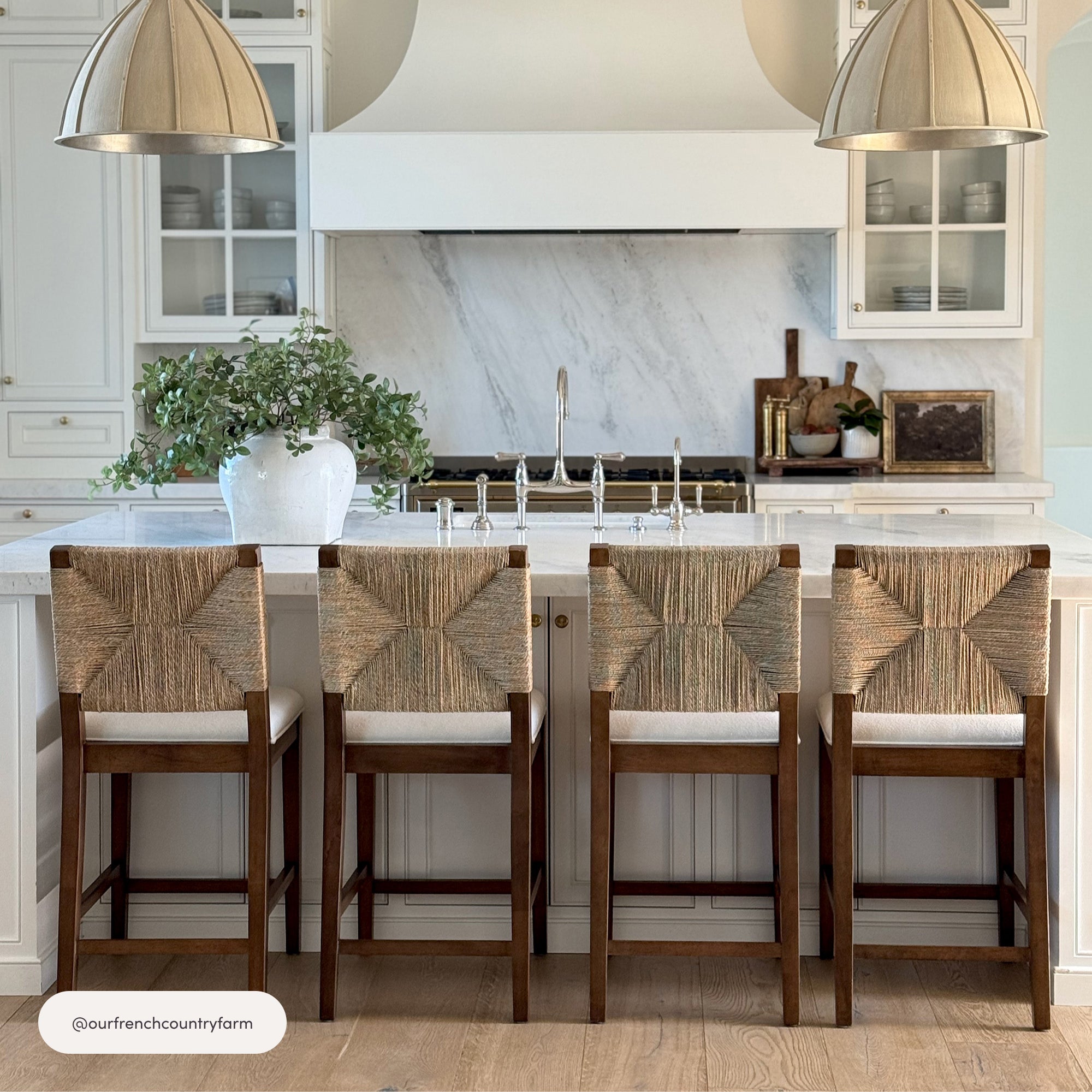 A bright, modern kitchen with a marble backsplash, four woven-back barstools at a white island, pendant lights above, and a large white vase with greenery on the countertop.
